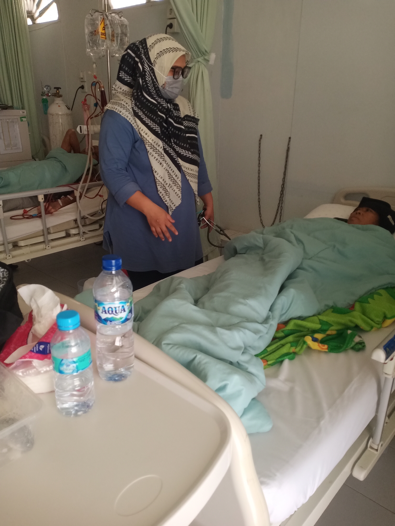 This image captures an interior scene within a medical facility located in Lhokseumawe, Indonesia, featuring two patients and one healthcare professional.
In the center-left of the frame, a female healthcare professional stands, attired in a blue long-sleeved top, a black and white patterned hijab, glasses, and a light blue surgical mask covering her mouth and nose. She holds a stethoscope in her left hand and appears to be observing the patient on the right.
To the left of the healthcare professional, a patient is lying on a hospital bed, predominantly covered by a light green blanket, with portions of their legs and a hand visible. This patient is connected to a medical system featuring red and blue tubing, indicative of a procedure such as dialysis. Adjacent to this bed, a medical monitor, two white oxygen cylinders, and an IV pole with multiple fluid bags, one distinctly labeled 'SIKO', are present.
To the right, a second patient is resting on a hospital bed, covered by a light blue blanket and an additional blanket with a green, yellow, and orange pattern. A dark head covering is visible on this patient's head.
In the immediate foreground, a white portable bedside table holds two clear plastic bottles of "Aqua" brand mineral water, one with a blue cap and the other with a lighter blue cap. Crumpled tissues or napkins and a clear plastic container are also on the table.
The room features light-colored walls and a tiled floor. Light green curtains are drawn across a window or opening on the left side, and another curtain is partially visible on the right. In the middle-right background, two metal chains hang vertically from the wall. A louvered window or vent is visible in the upper left portion of the wall.