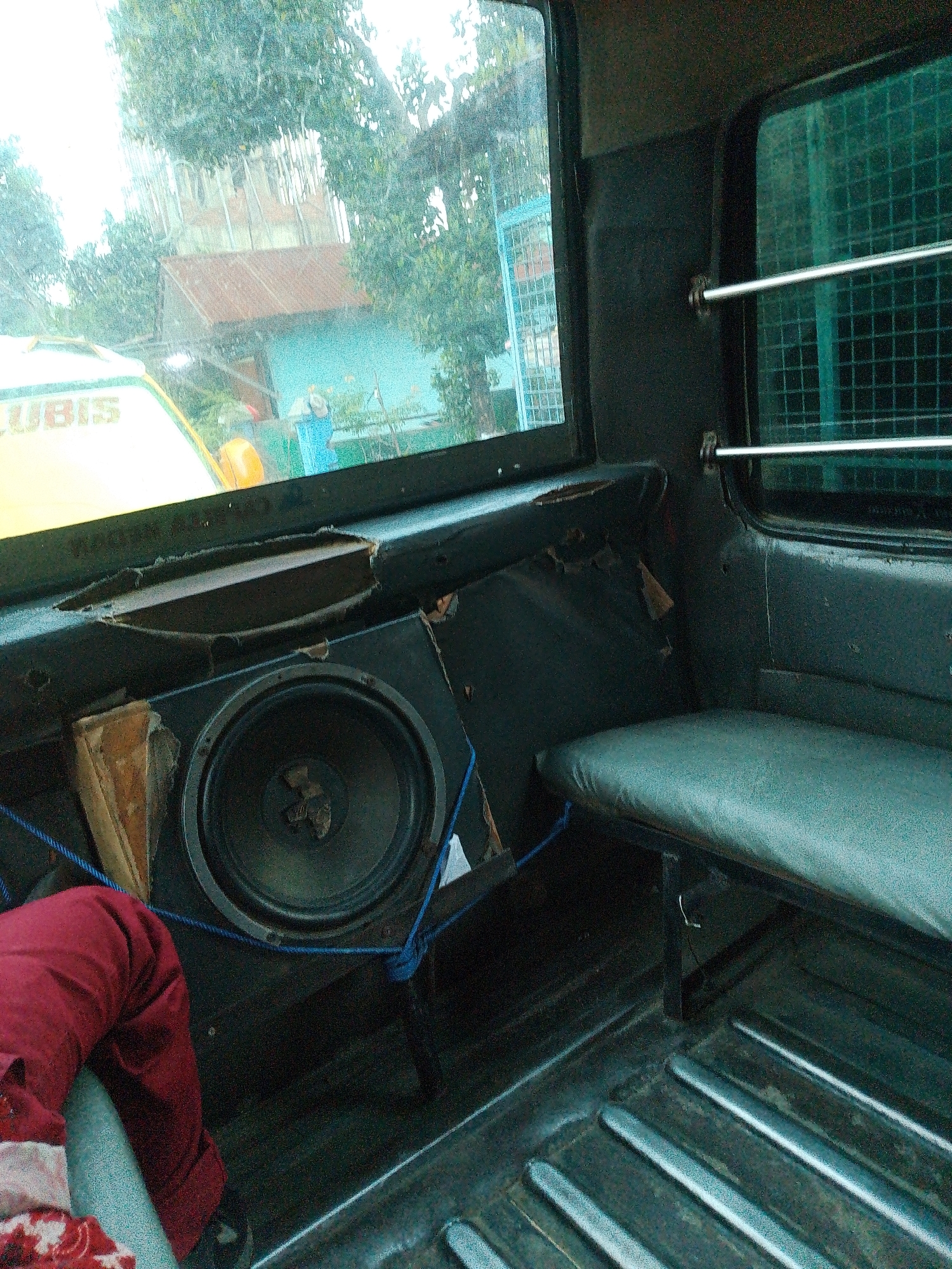 The image depicts the interior of a vehicle, likely a truck or van, seen from the perspective of a passenger seated on the left side. The dominant colors are dark grays and black, with a prominent gray upholstered bench seat on the right. A large, black speaker is visible in the lower left quadrant, mounted in a black enclosure. A piece of red fabric, possibly clothing, is draped over the edge of the seat in the foreground.
Through the vehicle's windows, an outdoor scene is visible, suggesting the vehicle is stationary. The exterior shows lush green trees and a structure with a reddish-brown corrugated metal roof and light blue walls. A partial view of a yellow vehicle with text, possibly "UBIS," is also visible to the left. The presence of raindrops on the window suggests it may be raining or has recently rained.
While the image provides a clear view of the vehicle's interior and the external environment, there are no discernible individuals or specific actions taking place within the frame. The overall condition of the vehicle's interior appears somewhat worn, with some visible damage to the paneling around the speaker. The scene suggests a typical, everyday setting within a residential or semi-urban area, potentially in Bandar Khalipah, Indonesia, given the context provided, but no definitive landmarks confirm this specific location.