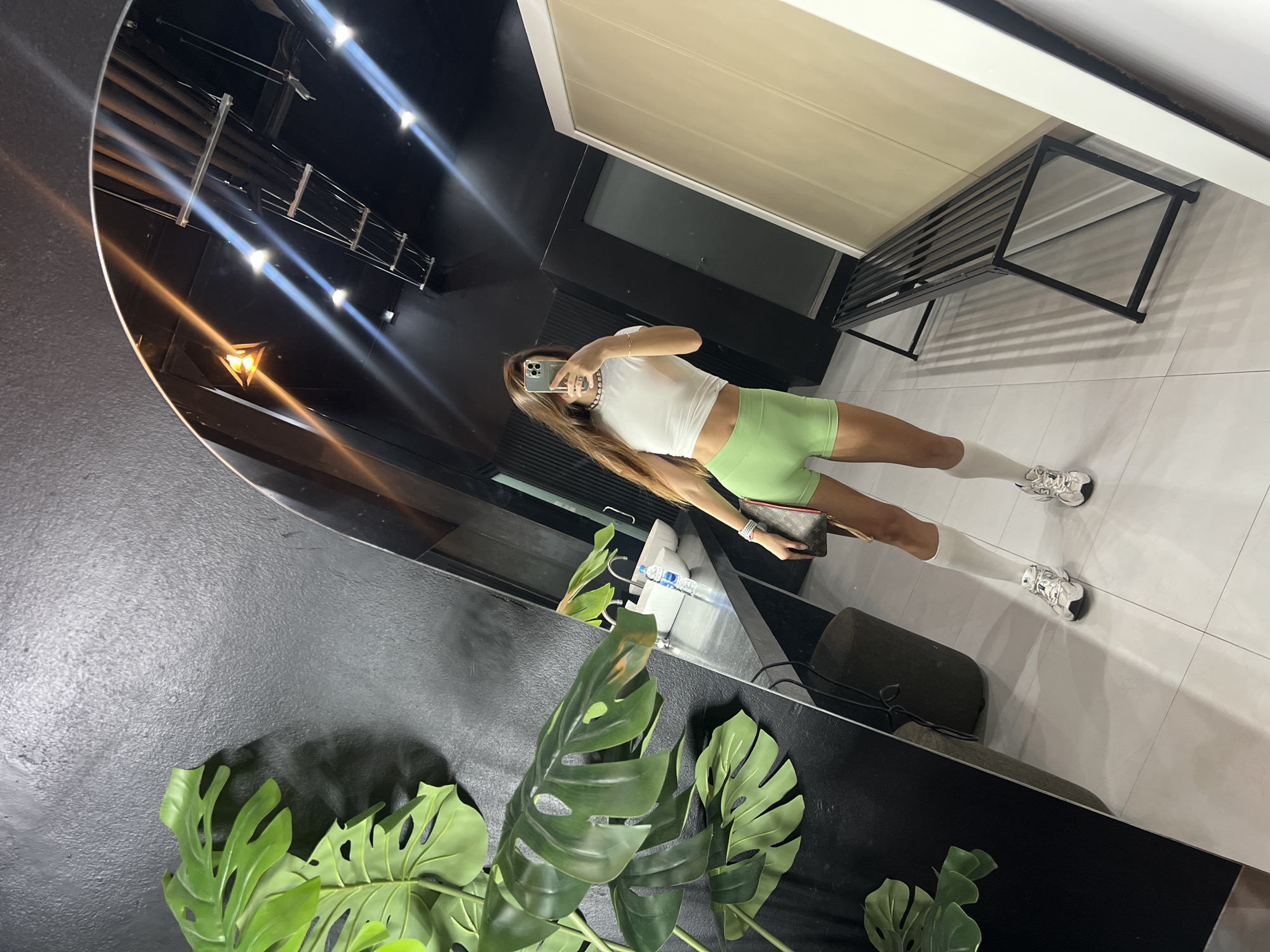 A young woman with long brown hair takes a mirror selfie in what appears to be a locker room or dressing area. She is wearing a white cropped t-shirt, light green shorts, white socks pulled up to her calves, and white athletic shoes. She is holding a brown designer handbag with a red trim and gold accents. In the background, reflected in the mirror, are sinks, a bench, a large artificial plant, and overhead lighting. The walls are dark gray, and the floor is tiled in a light color. The overall impression is one of a modern, minimalist aesthetic, likely in a gym, spa, or similar facility.