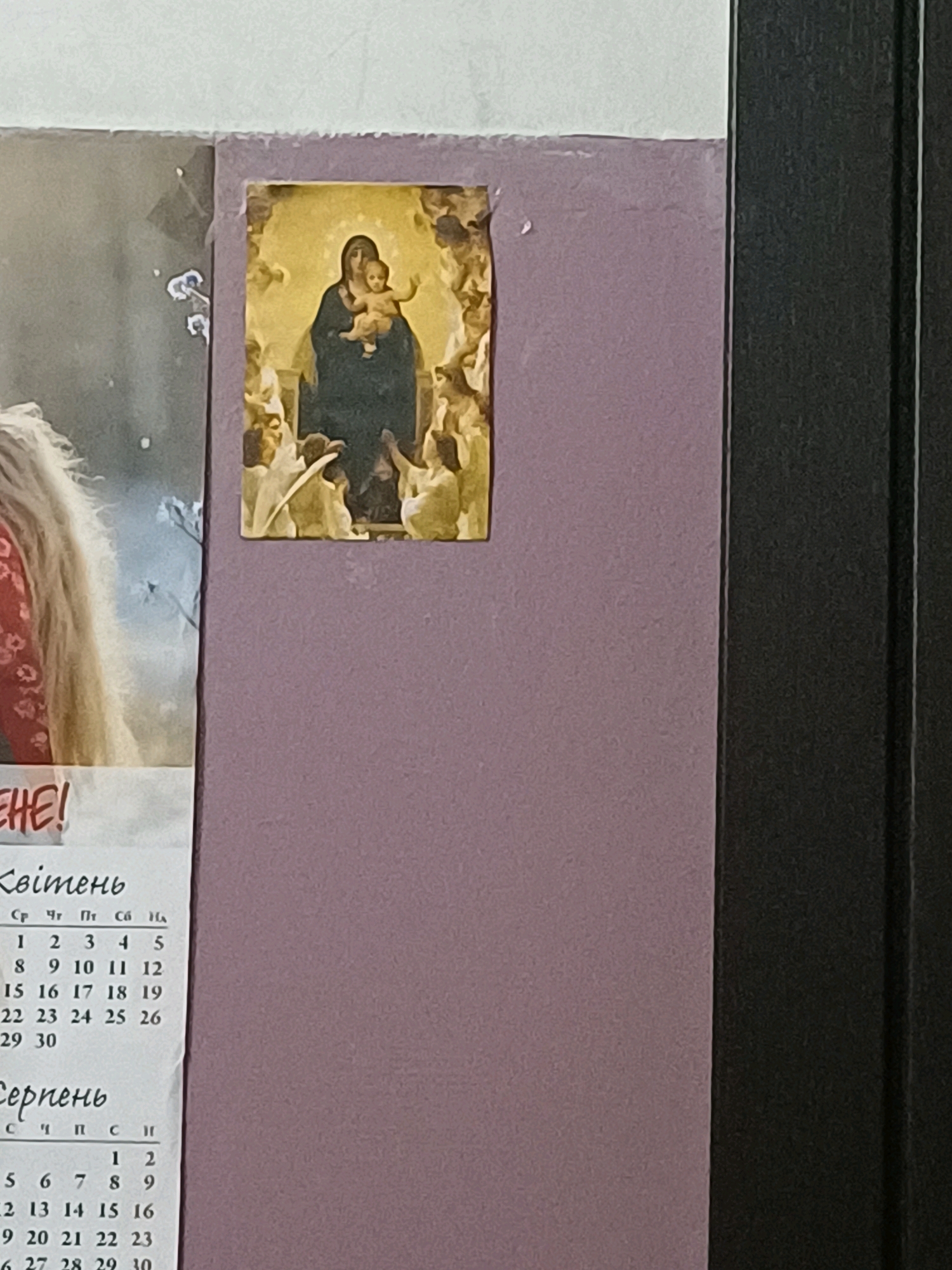 The image shows a close-up view of a wall with a small religious icon and a section of a calendar. The icon, depicting the Virgin Mary holding the infant Jesus surrounded by angels, is affixed to a purple wall. To the left of the icon, a portion of what appears to be a poster or calendar is visible, with the word "EHE!" clearly legible. Below this, a Ukrainian calendar for April ("Квітень") and August ("Серпень") is partially visible. The calendar lists days of the week in Ukrainian (Ср, Чт, Пт, Сб, Нд) and numerical dates. The scene appears to be indoors, possibly in a residential or public space in Kyiv, Ukraine, given the Ukrainian language on the calendar. A dark, vertical element, likely a door frame or furniture, is visible on the right side of the image.
