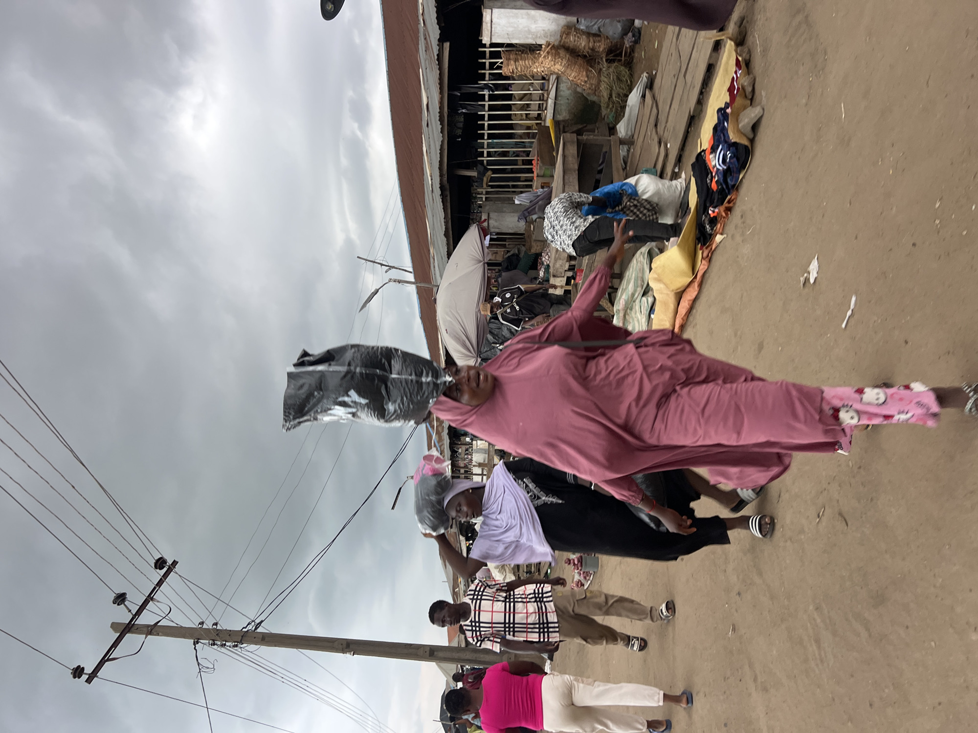 This is a medium-shot, eye-level, full-color, outdoor photograph taken at a market in Oko Amakom, Nigeria. The sky is overcast and gray, suggesting either a cloudy day or the onset of rain. A wooden utility pole with multiple insulators and wires is prominent on the left side of the frame, with some wires sagging. In the foreground, a woman wearing a rose-colored abaya and pink pants with white Hello Kitty patterns is walking towards the viewer, carrying a large black plastic bag on her head. She appears to be mid-stride, with her right arm extended forward. To her left, another woman wearing a black abaya and a lavender hijab is walking away from the viewer, also carrying a black plastic bag on her head. She is wearing white slides on her feet. A man in a white, pink, and black plaid shirt and beige cargo pants, wearing black and white slides, is walking behind her. Further left, a woman in a bright pink t-shirt and white pants is walking away from the viewer. In the background, a long, low building with a corrugated metal roof stretches across the frame. Several stalls with wooden tables and displays of goods, including woven baskets and what appear to be fabrics or clothing, are visible. Other individuals are present in the mid-ground and background, some appearing to be vendors or shoppers. The ground is dirt and appears dusty.