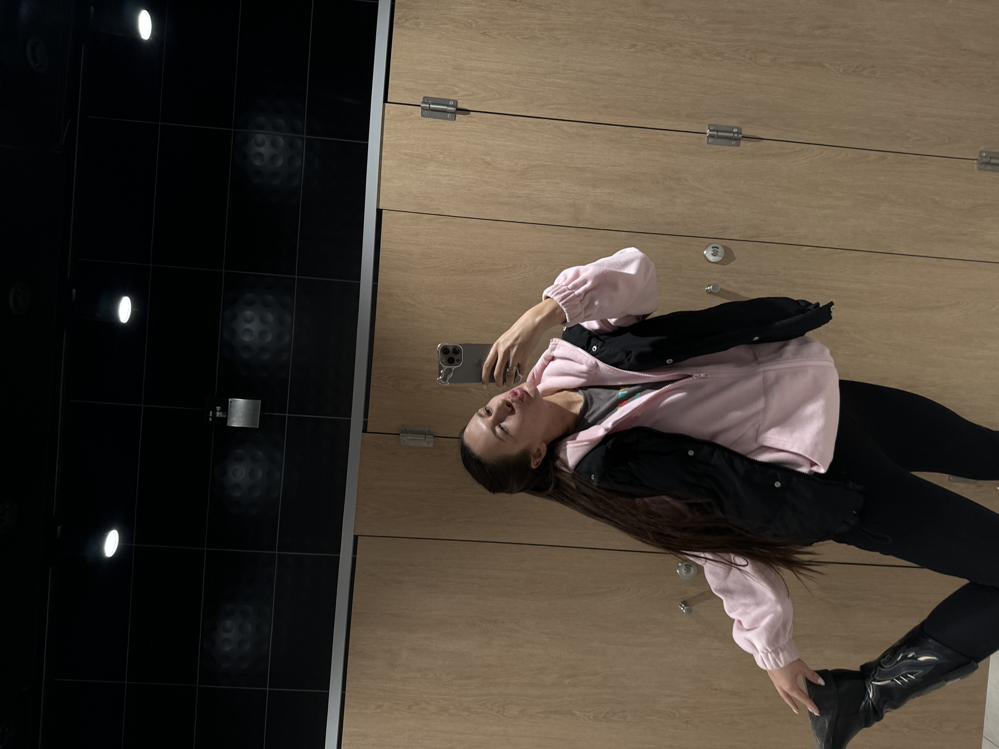 A woman is taking a selfie in what appears to be a locker room. She is wearing black pants, a pink jacket over a black jacket and black boots. She is holding a phone in her hand. The room has wooden lockers and black tiled walls. There is no indication of the location being Ivashevo, Russia.