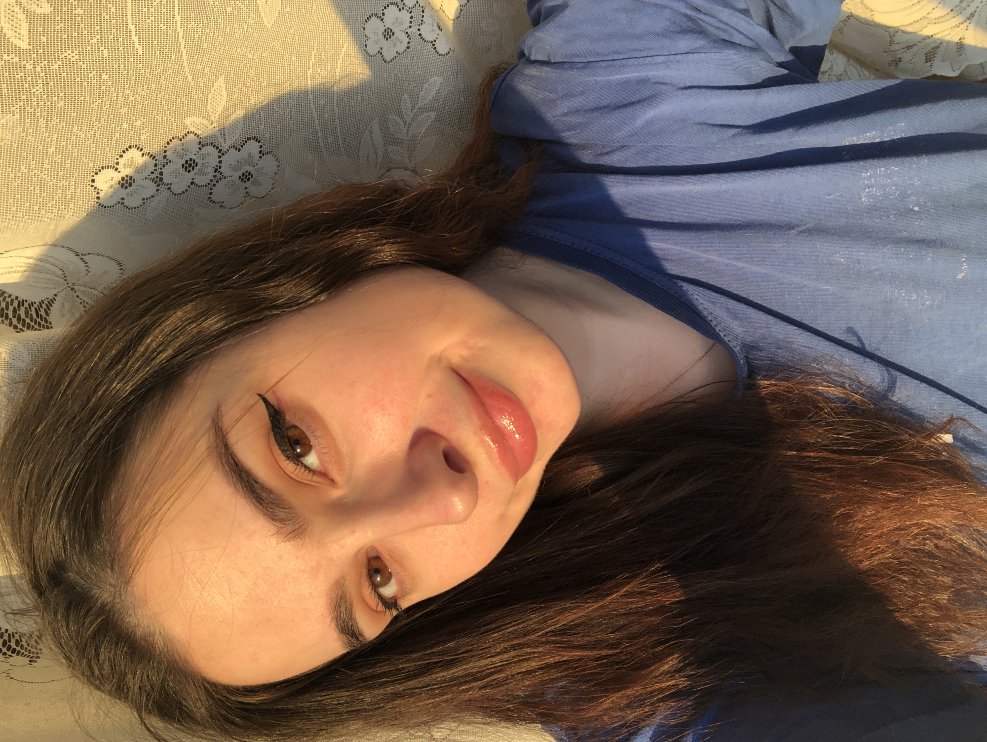 A young woman with long, brown hair lies on a light-colored surface with a floral pattern. She is wearing a blue top. Her eyes are brown and she is wearing eyeliner. The light source appears to be from the left side, creating shadows. The location is unknown; there is no indication it is Noviy, Russia.