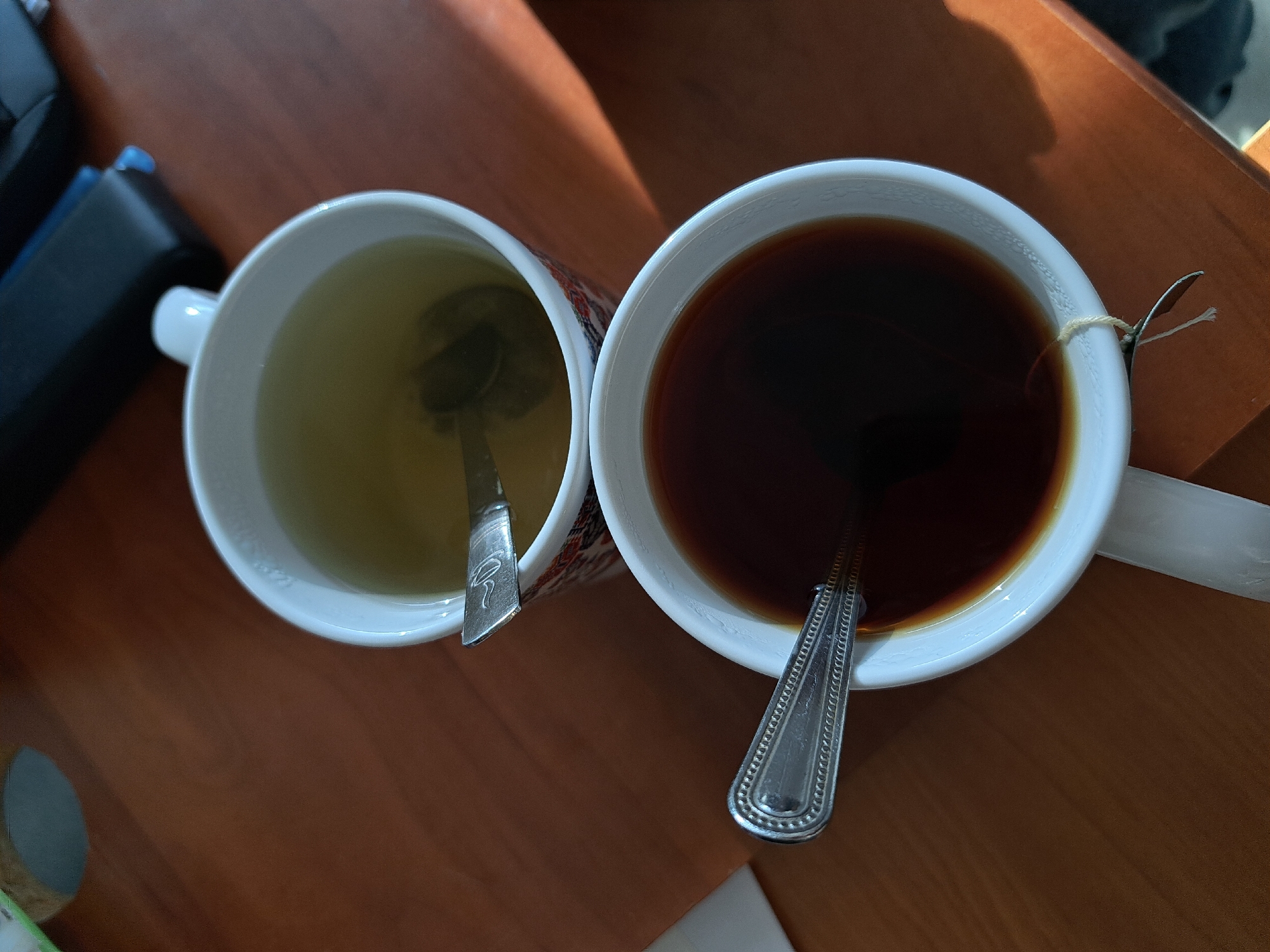 Two mugs of tea sit on a wooden surface. The mug on the left contains light-colored tea, possibly green tea, with a spoon inside. The mug on the right contains dark tea, likely black tea, also with a spoon inside and a teabag string dangling over the side. Part of a black object is visible to the left of the mugs. The location is Unknown, Unknown.