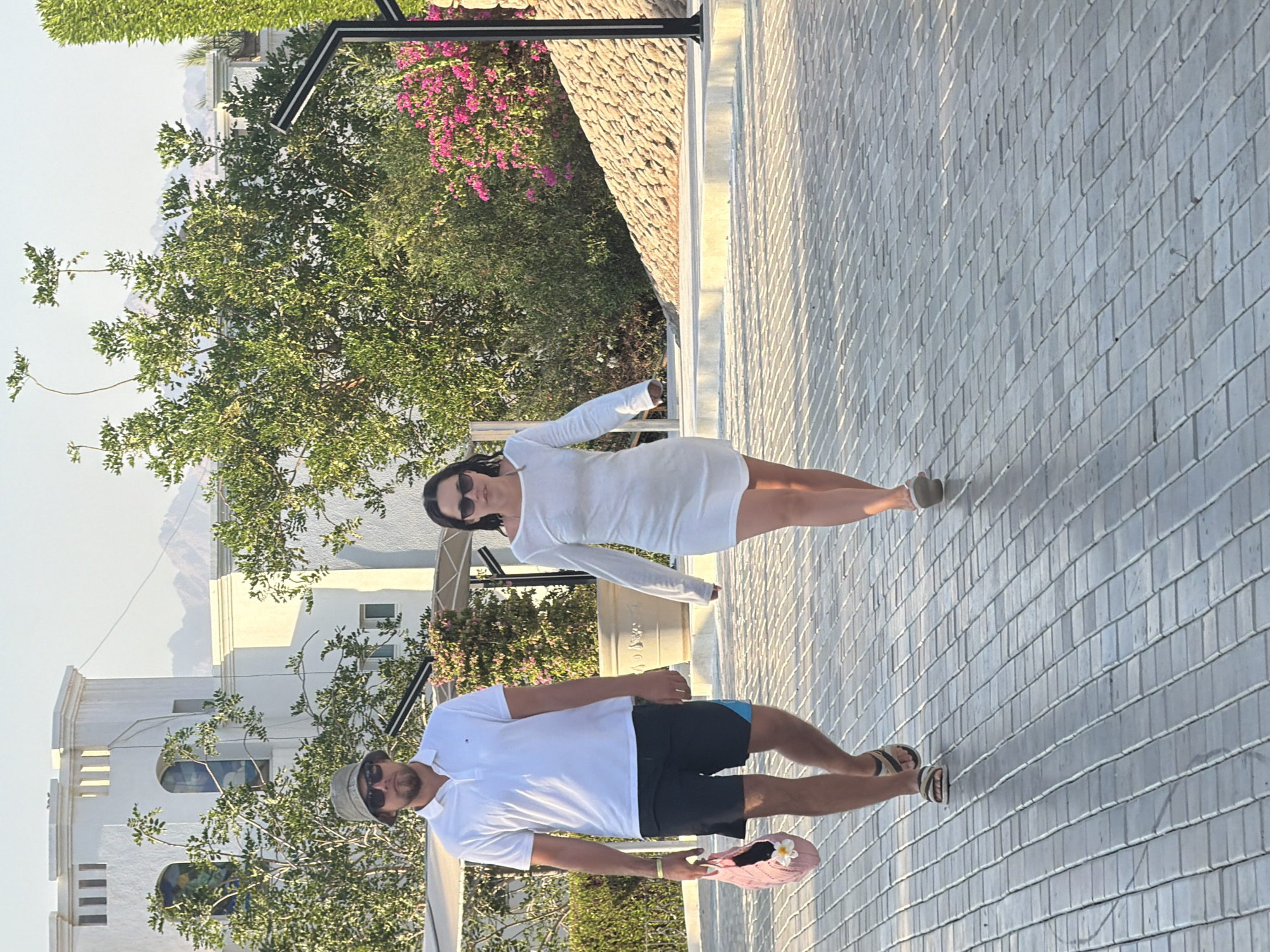 Two individuals are walking on a paved pathway in Sharm El Sheikh, Egypt. The person on the left is a male wearing a white polo shirt, black shorts with a blue accent, a bucket hat, sunglasses, and sandals. He is carrying a pink woven bag with a white flower embellishment. The person on the right is a female wearing a white long-sleeved dress and sunglasses. They are walking side-by-side. The background features a white building with arched windows, lush green trees, and pink flowering bushes. In the distance, mountains are visible under a clear sky. The pathway is made of grey rectangular pavers.