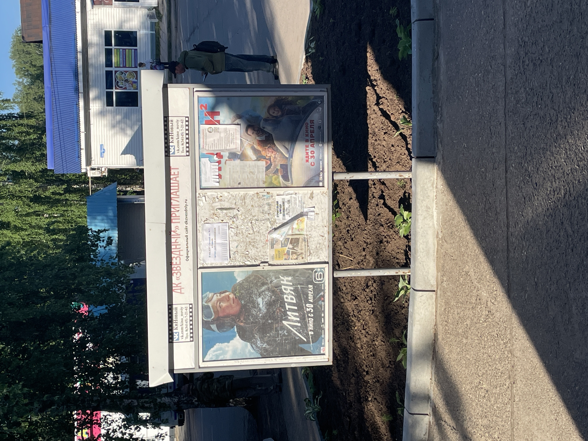 The image depicts an outdoor bulletin board in Noviy, Russia, displaying movie posters and information. The bulletin board is located on a sidewalk next to a street and is surrounded by a planter bed with dark soil and some young green plants. The board itself has a white header with text and logos, including "КЦ Киновый" and "ДК «ЗВЕЗДНЫЙ» ПРИГЛАШАЕТ". Below the header are two framed movie posters. The poster on the left features a portrait of a woman in a pilot's helmet with the title "ЛИТВЯК" and "В КИНО С 30 АПРЕЛЯ". The poster on the right displays a group of people in a car with the title "ЖДИТЕ В КИНО С 30 АПРЕЛЯ". There are also other flyers and papers attached to the bulletin board.
In the background, a white building with a blue corrugated metal roof is visible. A person in a green jacket and backpack stands on the sidewalk to the right of the bulletin board, looking at their phone. Another person in a camouflage uniform is partially visible on the left side of the image, standing near a tree. The overall environment appears to be a sunny day in a town or city setting.