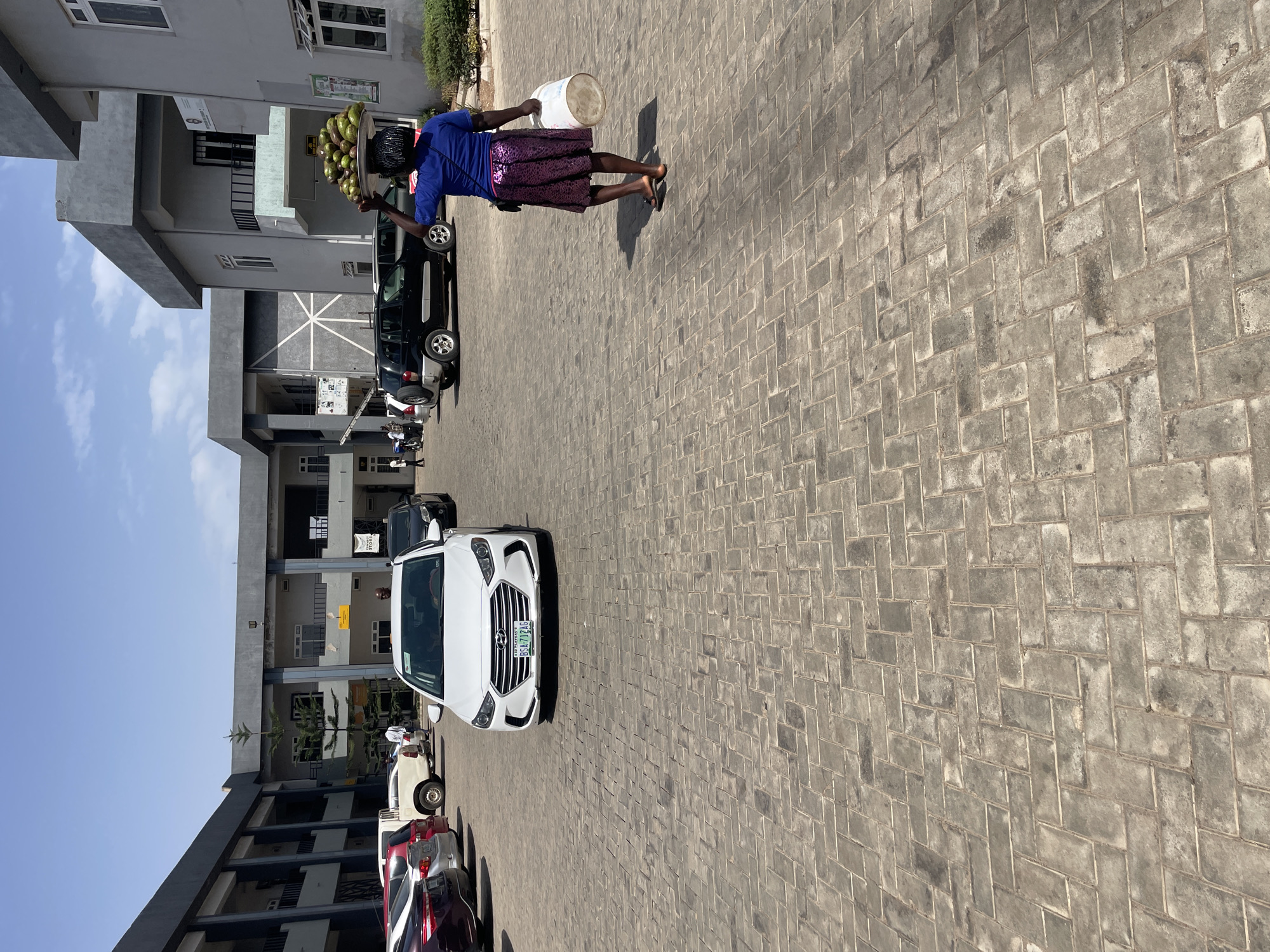The image captures an outdoor urban scene, likely a paved parking area or forecourt in Jos, Nigeria, under bright daylight. A woman is the primary subject, walking from right to left across the mid-ground. She is dressed in a blue short-sleeved top and a patterned purple skirt, wearing sandals. Expertly balanced on her head is a large, round metal tray laden with numerous green fruits, possibly avocados. In her left hand, she carries a white plastic bucket.
The ground is covered with grey interlocking concrete pavers, arranged in a herringbone pattern, showing some signs of wear and dirt. Distinct shadows cast on the pavement indicate strong sunlight, suggesting it is mid-morning or early afternoon. The sky is clear and blue.
Several vehicles are parked in the scene. A white Hyundai sedan is prominent in the foreground to the left of the woman, facing the viewer. Its Nigerian license plate, "BSN 721 LG," is clearly visible. Further back and to the left, a dark SUV and other cars are parked.
In the background, multi-story buildings with grey facades and multiple windows line the area. These appear to be commercial or residential structures. Some signs are visible on the buildings; one yellow sign is unreadable, while another white sign on a building to the far right appears to partially read "STONE" or similar text. The overall scene depicts a typical busy street or market area, where commerce and daily life unfold.