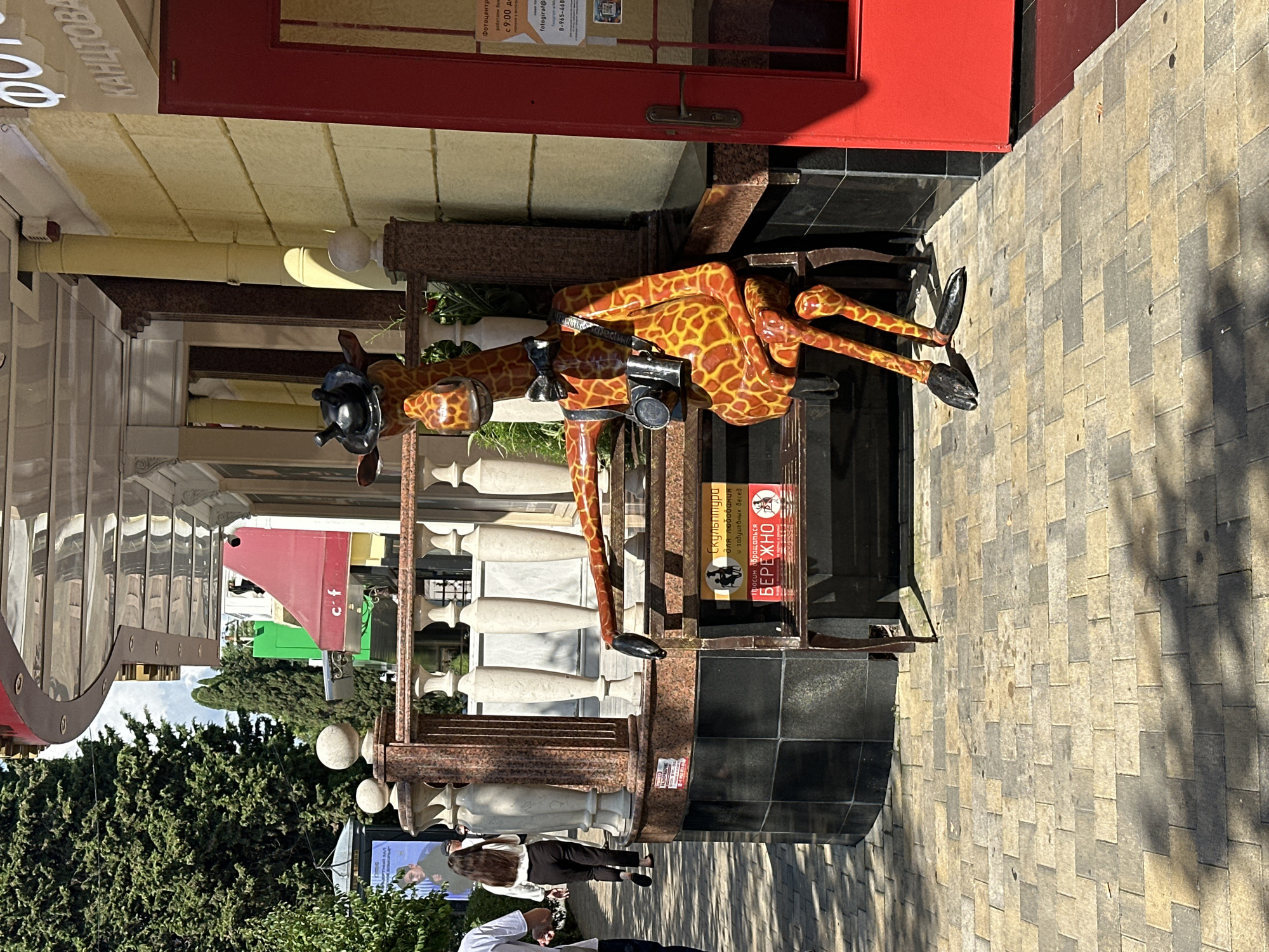 An outdoor scene in Sochi, Russia, shows a life-sized, decorative giraffe sculpture with a camera around its neck, sitting on a bench in front of a building with a red door and yellow facade. The giraffe is wearing a black hat and bow tie. Two signs are visible on the bench behind the giraffe. One sign, in Russian, has text that translates to "Sculpture for visitors and the people who came to visit". The other sign is red with a white border and has an image of a person on it with text that says "BE CAREFUL". The ground is paved with interlocking bricks, and there are trees and architectural elements of a building in the background.