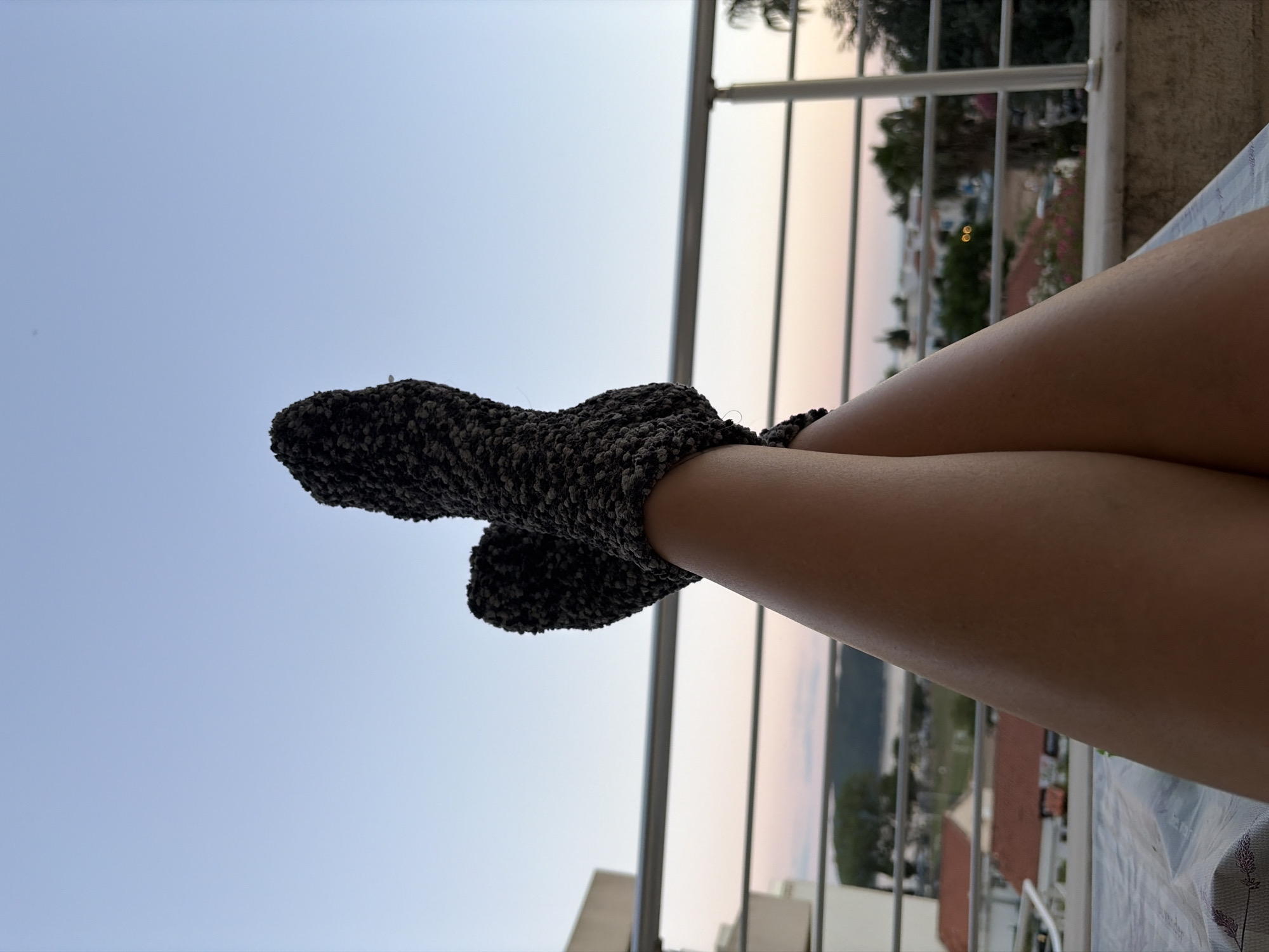 The image captures a low-angle perspective of a person's legs, clad in fuzzy, dark-colored socks, resting on what appears to be a sun lounger. The legs are crossed at the knees, with the feet pointing upwards towards a clear sky that transitions into hues of pink and orange near the horizon, suggesting it is either dawn or dusk.
In the background, a balcony railing with horizontal silver bars runs across the frame, separating the viewer from the scene beyond. Below the railing, a coastal town is visible. Buildings with red-tiled roofs and white facades are scattered throughout the landscape. Lush green trees, including palm trees, dot the area, and a body of water, possibly the Adriatic Sea, can be seen in the distance, with hills or mountains forming a backdrop. The overall setting appears to be a residential area or a resort in Budva, Montenegro, likely a balcony or terrace overlooking the town and coastline. The lighting indicates an outdoor setting during the twilight hours.