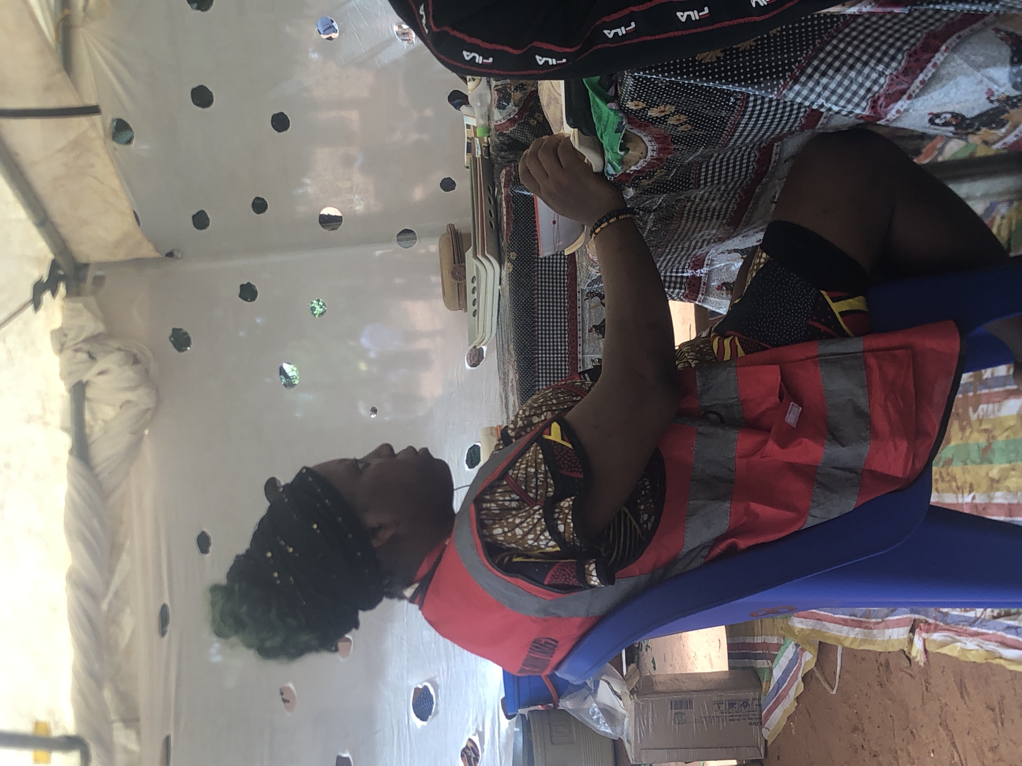A person with dark skin sits on a blue plastic chair inside a structure covered in white plastic sheeting with circular holes. The person wears a red and gray striped safety vest over a patterned, multicolored shirt. The individual has black hair gathered on top of their head. At the top of the image, another person is partially visible, wearing a black top with "FILA" branding. A checkerboard pattern is also visible in their clothing. A table is nearby with various objects, including a container and a straw hat. The structure appears to be a temporary enclosure, as evidenced by the sheeting. The location is Nteje, Nigeria.