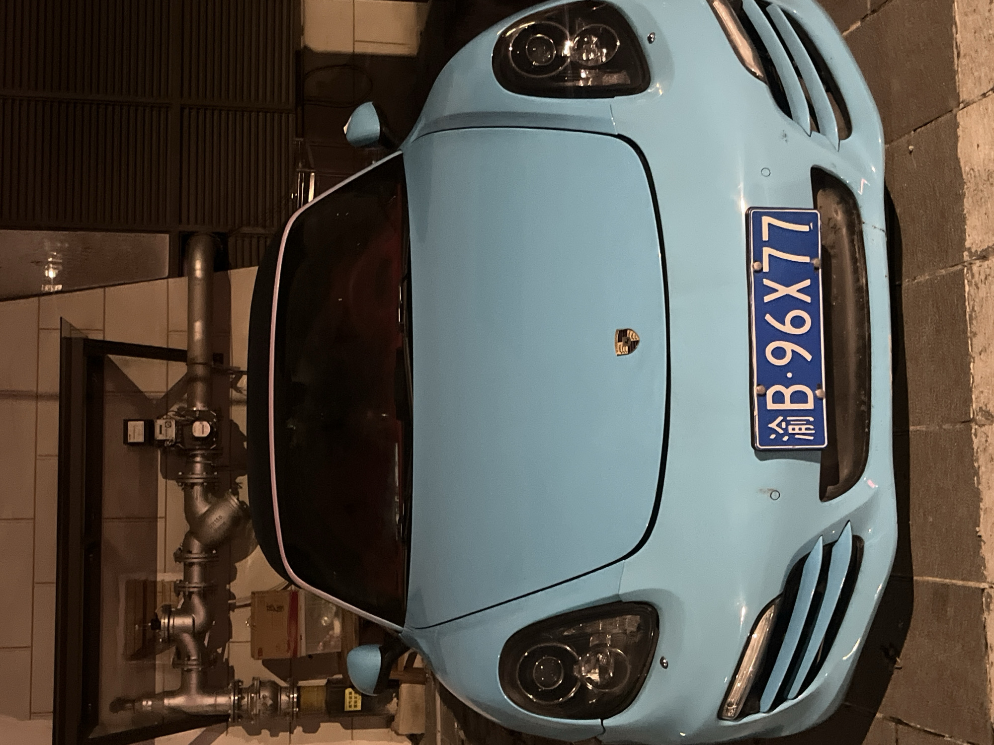 A light blue Porsche Cayman is parked in front of a building with exposed pipes and ventilation grates. The car's license plate reads "渝B·96X77." The scene appears to be outdoors at night, with artificial lighting illuminating the vehicle and its surroundings. The ground beneath the car is paved with interlocking bricks. There are no people visible in the image. The background consists of a brick wall and what appears to be a dark-colored louvered vent. This location is identified as Jiulongpo District, China.