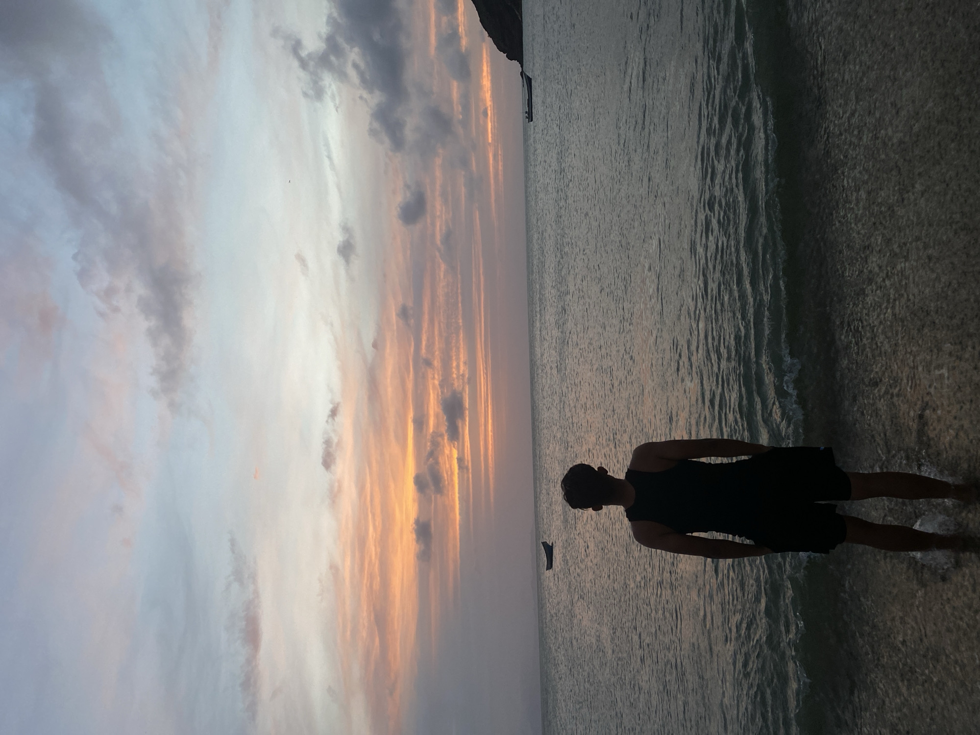 A vertical photograph shows a person standing in water at sunset. The person is facing the right, silhouetted against the water. The person is wearing a dark, sleeveless shirt and dark shorts. The water reflects the sky, which is striated with orange, pink, and gray clouds. A dark shape, possibly a boat, is visible in the distance. The location is Juan Griego, Venezuela.