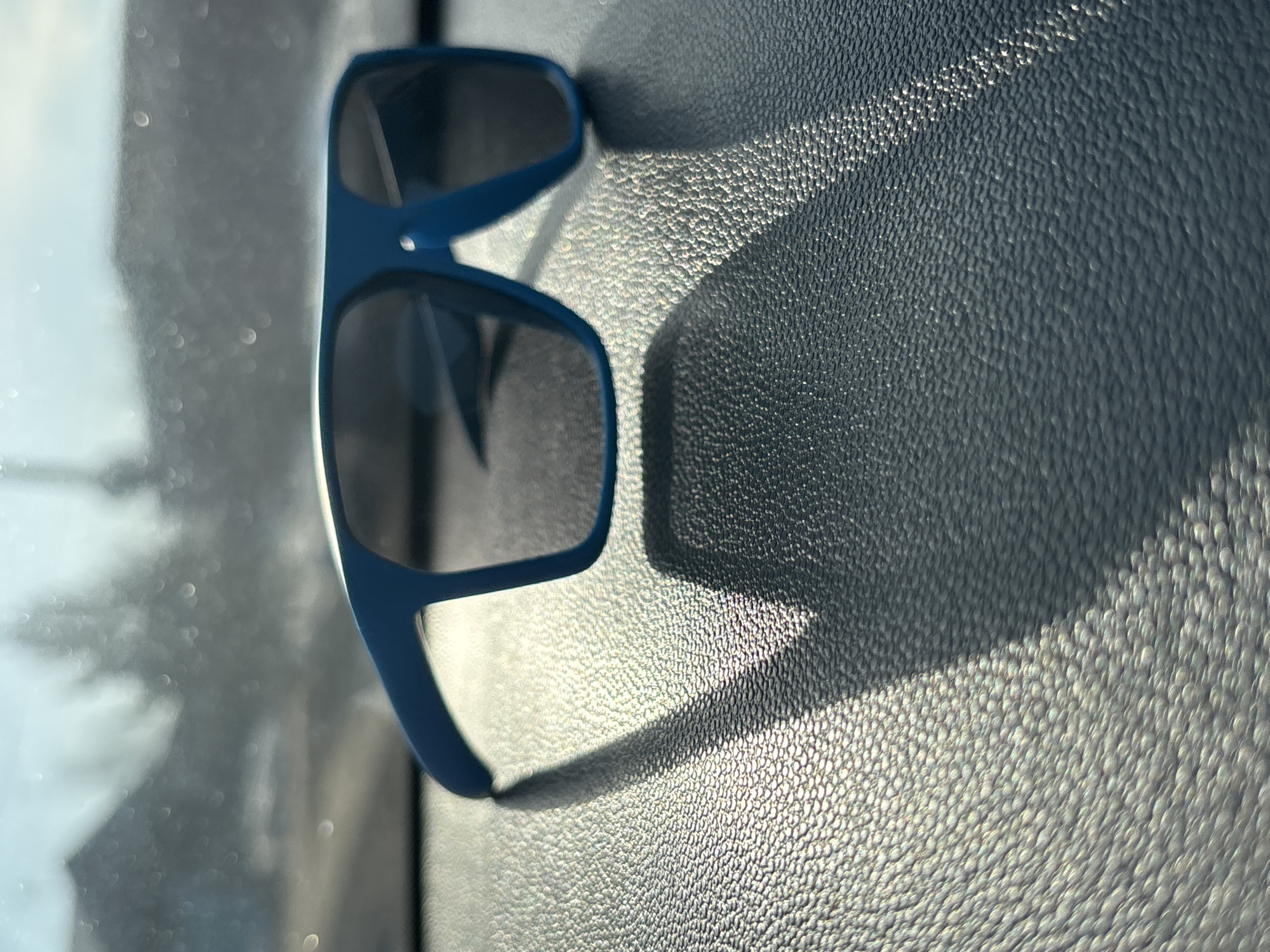 A pair of blue sunglasses lies on a textured surface, casting a shadow to the right. The surface appears to be a rough, pebbled material, possibly the dashboard of a vehicle. A blurred area to the left suggests a window with outside light. The location in the city of Chaikovsky, Russia is not determinable from the image alone.