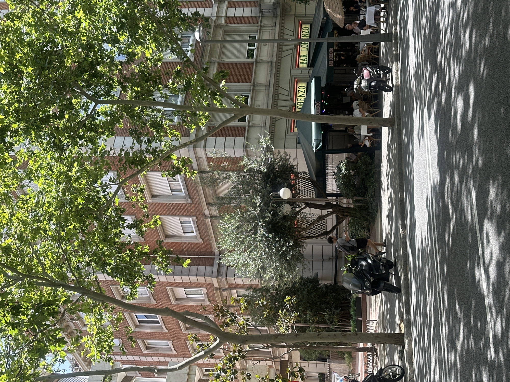 The image depicts a street scene in Barcelona, Spain, likely in the Eixample district due to the architectural style of the building and the presence of plane trees. A multi-story brick building with large windows and decorative stone accents dominates the background. In front of the building, a cafe or bar named "BAR LORENZO" is visible with patrons seated at tables under a green awning. A sidewalk runs alongside the street, shaded by mature trees casting dappled shadows on the asphalt. A black scooter is parked on the sidewalk, and a person wearing a gray t-shirt and dark shorts is walking away from it. Another scooter is partially visible on the far left of the frame. The overall atmosphere is one of a sunny day in an urban environment.