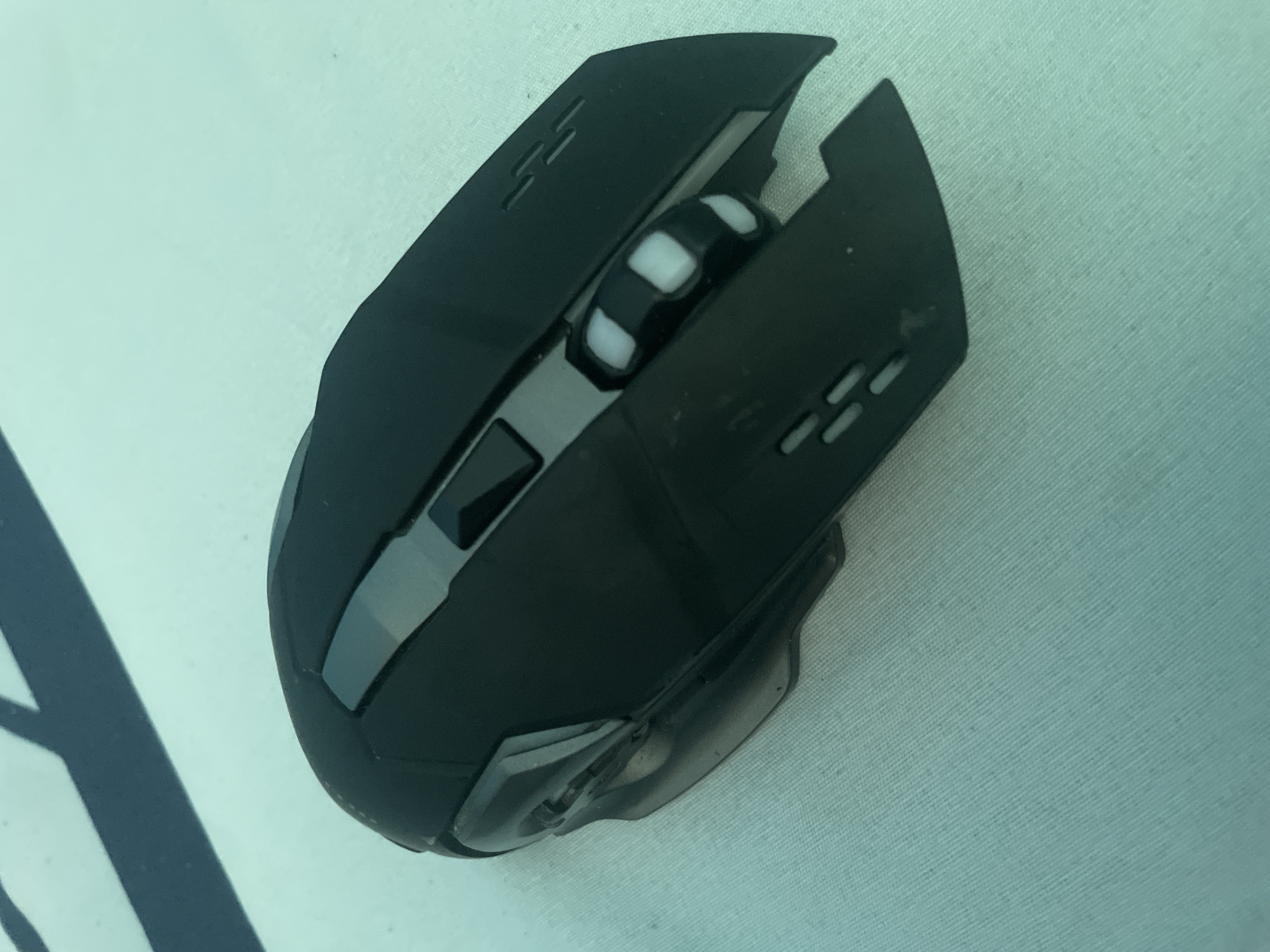 The image displays a close-up, overhead view of a black gaming mouse positioned on a light blue textured surface, possibly a mousepad or fabric. The mouse has a sleek, ergonomic design with several distinct features. The scroll wheel is prominently visible, featuring a black rubber grip with alternating white squares, giving it a checkered appearance. Two prominent buttons are located on the top surface of the mouse, one slightly to the left and another more centered. The left and right click buttons are integrated into the main body of the mouse.
The mouse has angular design elements, particularly on its sides, with what appear to be vents or decorative lines on the upper surfaces. A translucent or clear plastic section is visible towards the rear of the mouse, possibly housing lighting elements or the optical sensor. The surface beneath the mouse has a subtle ribbed texture and is a consistent, soft blue hue, suggesting an indoor setting. There is a dark, angled shadow cast by an unseen object on the left side of the frame, partially obscuring the edge of the mousepad. No people or explicit activities are depicted; the focus is solely on the object. No visible text is present on the mouse or its surroundings. The lighting appears even, indicating artificial light or diffused natural light. The overall impression is that of a product shot or a still life composition focused on the computer accessory. The location context provided (Kołobrzeg, Poland) does not offer any direct visual cues within the image itself.