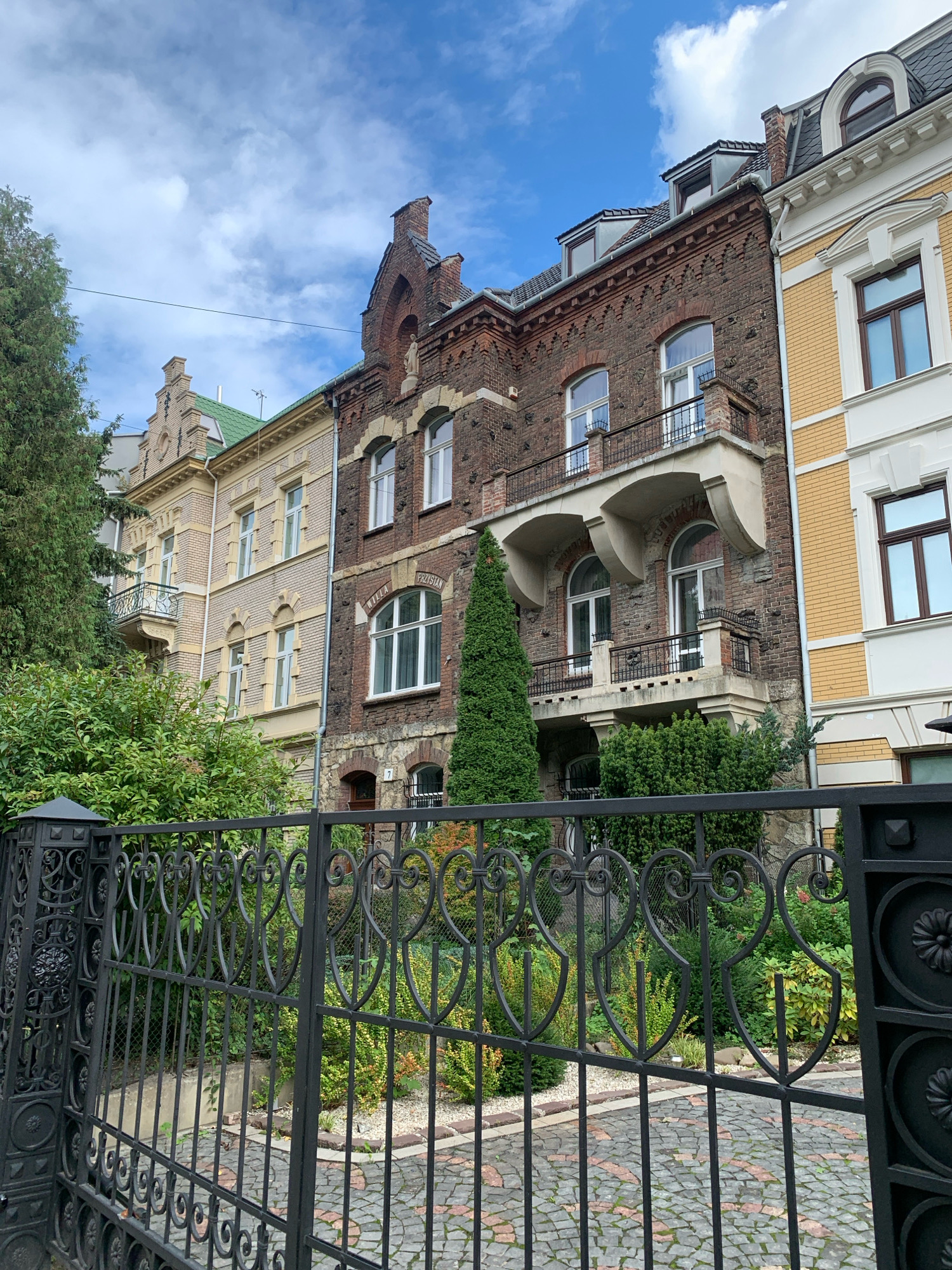 The image depicts a street scene in Lviv, Ukraine, focusing on several attached historic buildings and an ornate black metal gate in the foreground. The buildings, likely residential, display architectural styles from different eras, featuring brickwork, ornate facades, arched windows, and balconies. One building is a dark brick structure with decorative brickwork above the windows and arched windows on the upper floors. Adjacent to it is a lighter-colored building with decorative stonework and arched windows. Further down the street, another building with a light yellow facade and decorative white elements is visible. The sky is partly cloudy with a bright blue expanse. Behind the gate, a garden with various green shrubs and trees is visible, with a cobblestone path leading further into the property. The gate itself is intricately designed with scrolling metalwork and decorative elements. The scene suggests a well-maintained urban environment with a blend of historical architecture and lush greenery.
