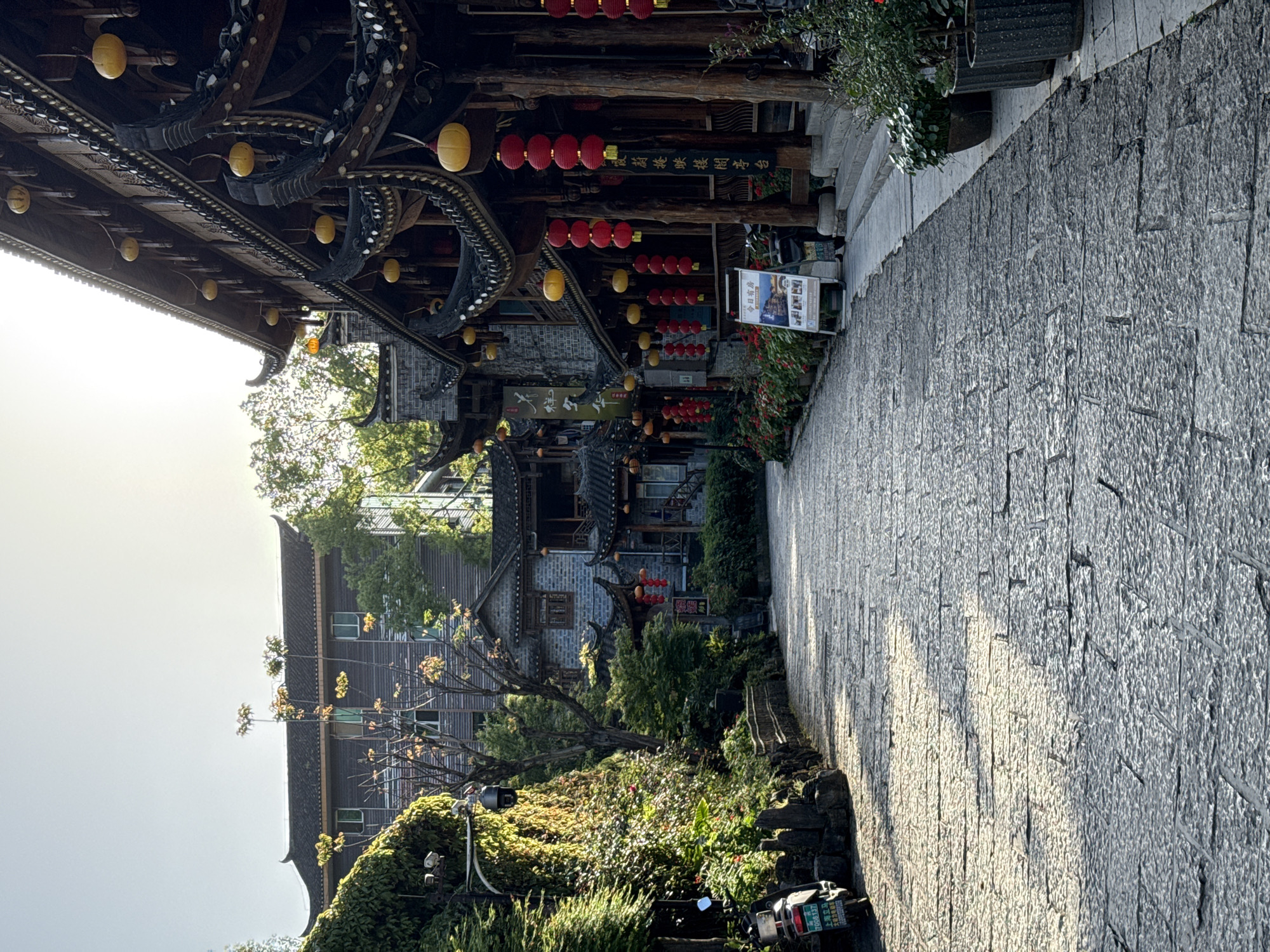 The image captures a street in Furong, China, characterized by traditional architectural elements. The buildings lining the street feature intricate wooden structures adorned with yellow and red lanterns. The street itself is paved with stone. Lush greenery is visible, adding to the scene's ambience. A scooter is parked on the side of the street. The image suggests a blend of historical charm and modern-day functionality in the city.