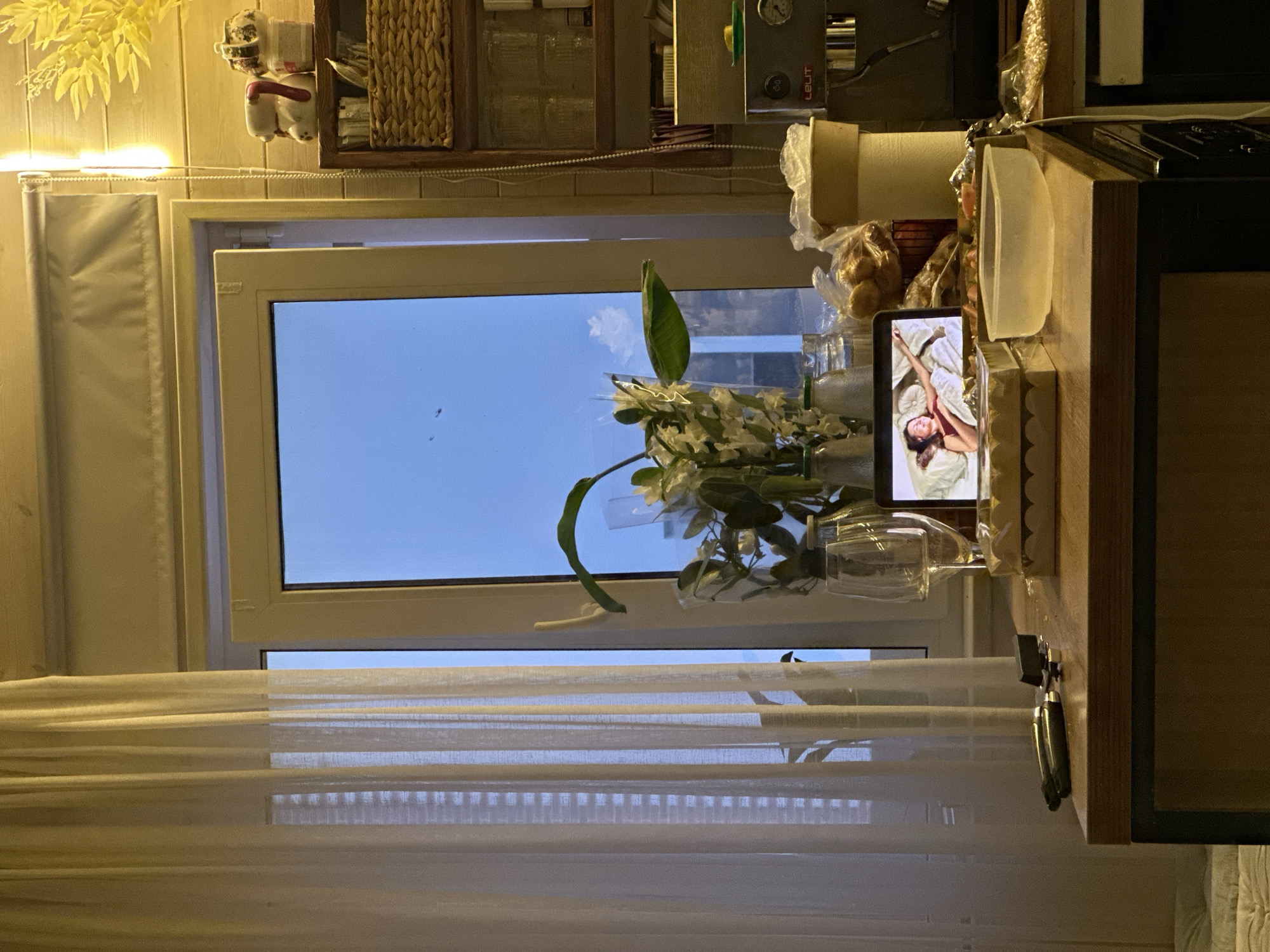 The image depicts a kitchen counter with a laptop displaying a woman in bed. To the left, sheer white curtains cover a window. A bouquet of white flowers in a vase sits on the counter, accompanied by glassware and bottles. In the background, a coffee machine is visible. The scene suggests a domestic setting, possibly a kitchen, with a focus on personal space and relaxation. The presence of the laptop and the woman's image implies digital engagement or entertainment. The overall ambiance is calm and intimate, with soft lighting and a hint of evening or dusk outside the window. The location, Тумілавіцкі сельскі Савет, Belarus, is not explicitly visible in the image but is provided as contextual information.