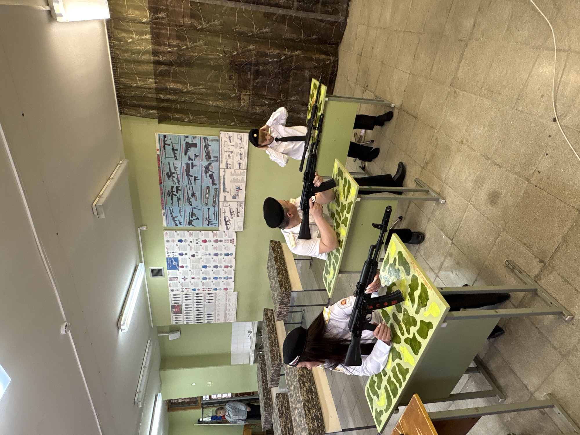 In the city of Markovskii, Russia, a classroom-like setting features three individuals seated at desks, each holding what appear to be rifles. The individuals are dressed in white shirts, black ties, and black caps, suggesting a uniform. The desks are decorated with a green and yellow camouflage pattern. Educational posters displaying firearms and military insignia adorn the wall. The room is lit by fluorescent lights. In the background, there is a partially visible individual behind a display case.