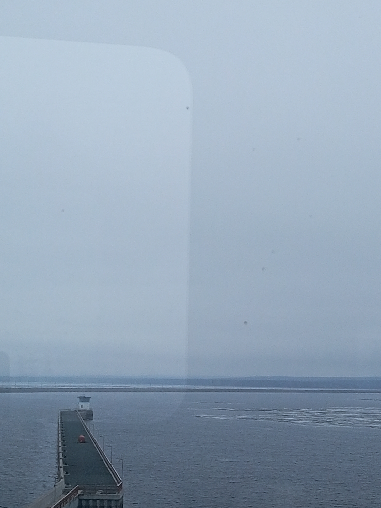 The image depicts a pier extending into a body of water, possibly a reservoir or large lake. At the end of the pier stands a small, white lighthouse-like structure. An orange buoy or marker is visible along the pier's surface. The water has some patches of lighter-colored material, possibly ice or sediment. In the background, a low-lying landmass is visible, obscured by haze or fog. The sky is overcast and gray. The scene appears to be viewed through a window, with some reflections or imperfections visible in the glass. Based on the context of Chaikovsky, Russia, this likely depicts the pier and light at the end of the Votkinsk Reservoir.