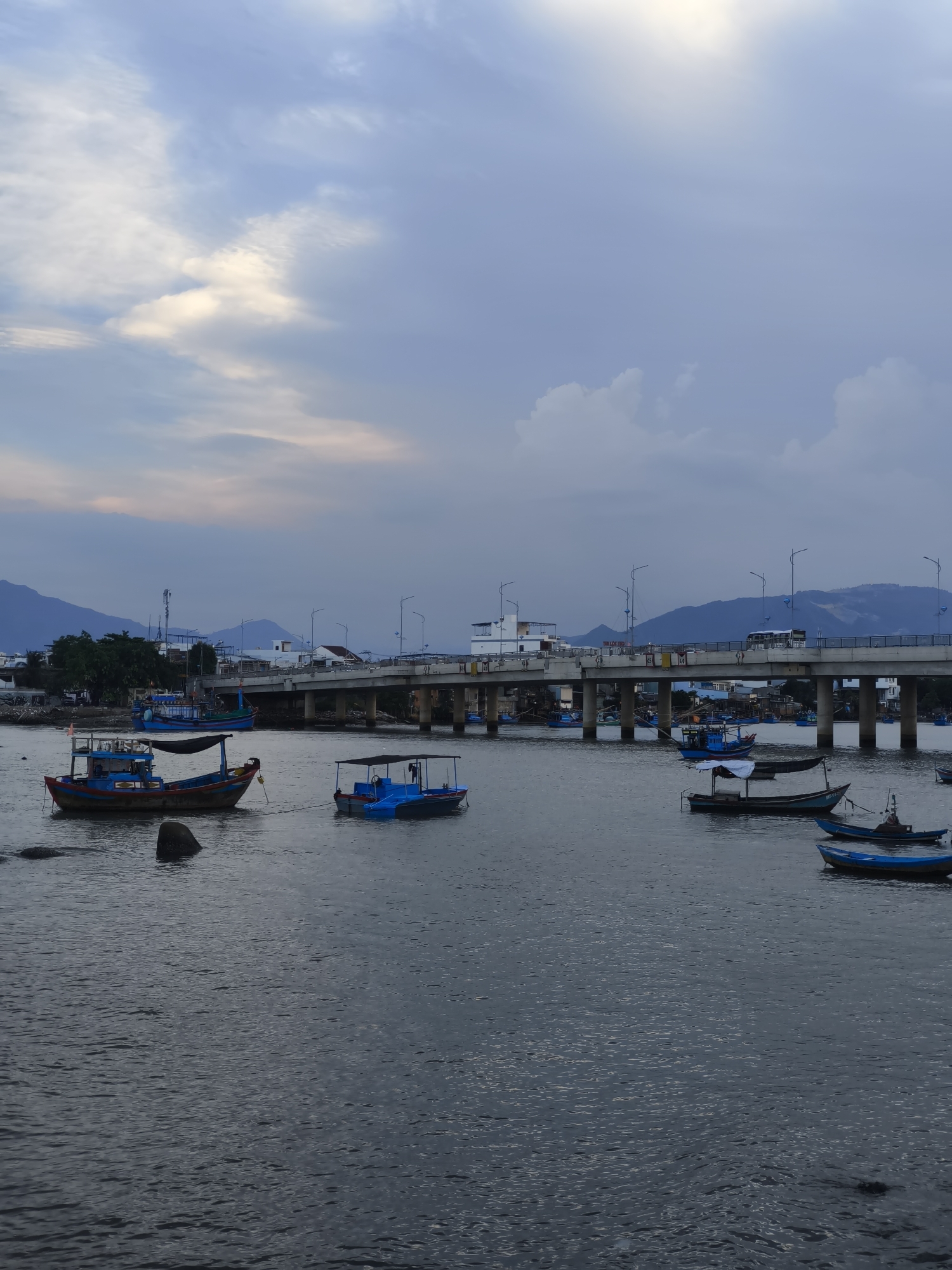 A wide shot captures a serene river scene in Bac Nha Trang Ward, Vietnam, likely during the late afternoon or early evening. The sky is a blend of soft blues and grays, with scattered clouds catching the fading sunlight. A long concrete bridge with numerous supporting pillars stretches across the water in the middle ground, with vehicles and streetlights visible on its surface. Several boats of various sizes and colors are moored or anchored in the river. Some are simple fishing boats with blue hulls and cabins, while others are smaller and more basic. The riverbanks in the distance are lined with buildings and lush green trees, with mountains forming a hazy backdrop. The water's surface is calm, with gentle ripples reflecting the muted light. The overall atmosphere is peaceful and tranquil.
