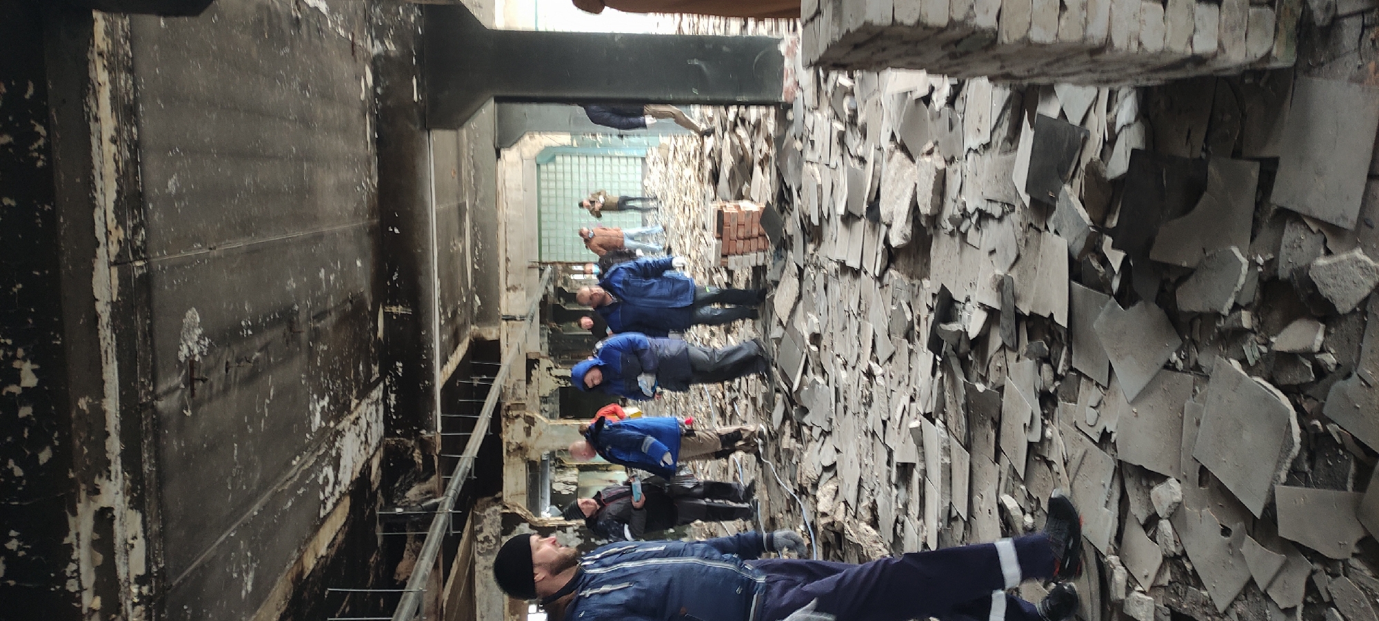 The image, oriented vertically and requiring rotation for correct viewing, captures an interior scene within a large, damaged, and derelict building, likely industrial or commercial. The floor is extensively covered with debris, consisting primarily of broken grey tiles and fragmented concrete. In the middle ground, stacks of red bricks are also visible among the rubble.
Numerous individuals, predominantly men, are present throughout the space, appearing to walk through or observe the damaged environment. They are dressed in practical, warm clothing such as jackets, some with hoods, and hats, suggesting a cool indoor temperature. Their postures and gazes indicate they are surveying the surroundings, looking at the debris, the structural elements, and towards different parts of the building.
To the left, a large, dark wall shows significant signs of wear and damage, including peeling paint, discoloration, and streaks, possibly from water damage or soot. Structural beams and columns are visible, contributing to the impression of a robust, possibly industrial, construction now in disrepair. In the background, a wall featuring translucent glass blocks allows diffused light into the space, indicating daytime. The overall atmosphere suggests an inspection, urban exploration, or a tour of a site that has suffered significant damage or long-term neglect.
No discernable text is visible on any surfaces, clothing, or objects within the image.