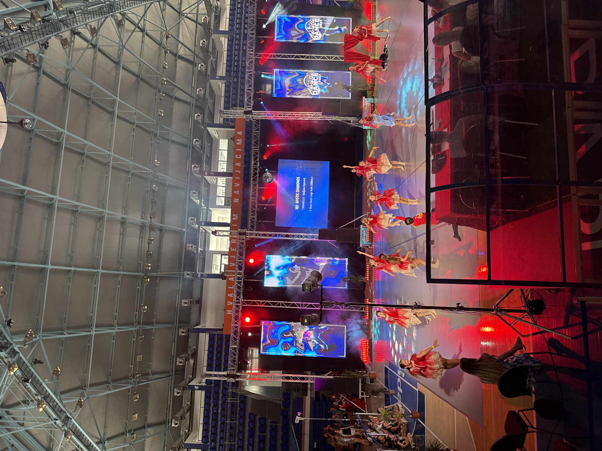 The image depicts a dance competition taking place in the Topolica Sports Hall in Bar, Montenegro. A group of young dancers, dressed in red and white costumes, are performing on a stage illuminated by colorful lights and large LED screens. The screens display abstract patterns and event logos, one of which reads "Adriatic Dance Fest." Several rows of blue stadium seating are visible in the background, mostly empty. In the foreground, a few spectators are seated, and a woman is sitting on a chair observing the performance. The overall scene suggests a lively event with a focus on youth dance performance.