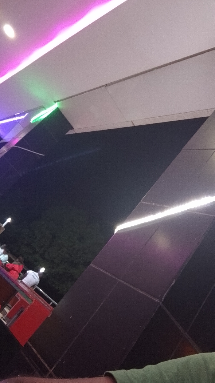 An angular nighttime shot features modern architecture adorned with colorful LED lighting. White, pink, and green lights illuminate various sections of the building's exterior. A glimpse of a red structure with people on it is visible in the foreground, suggesting an amusement park setting. Tiles and dark panels form geometric shapes, contributing to the contemporary design.