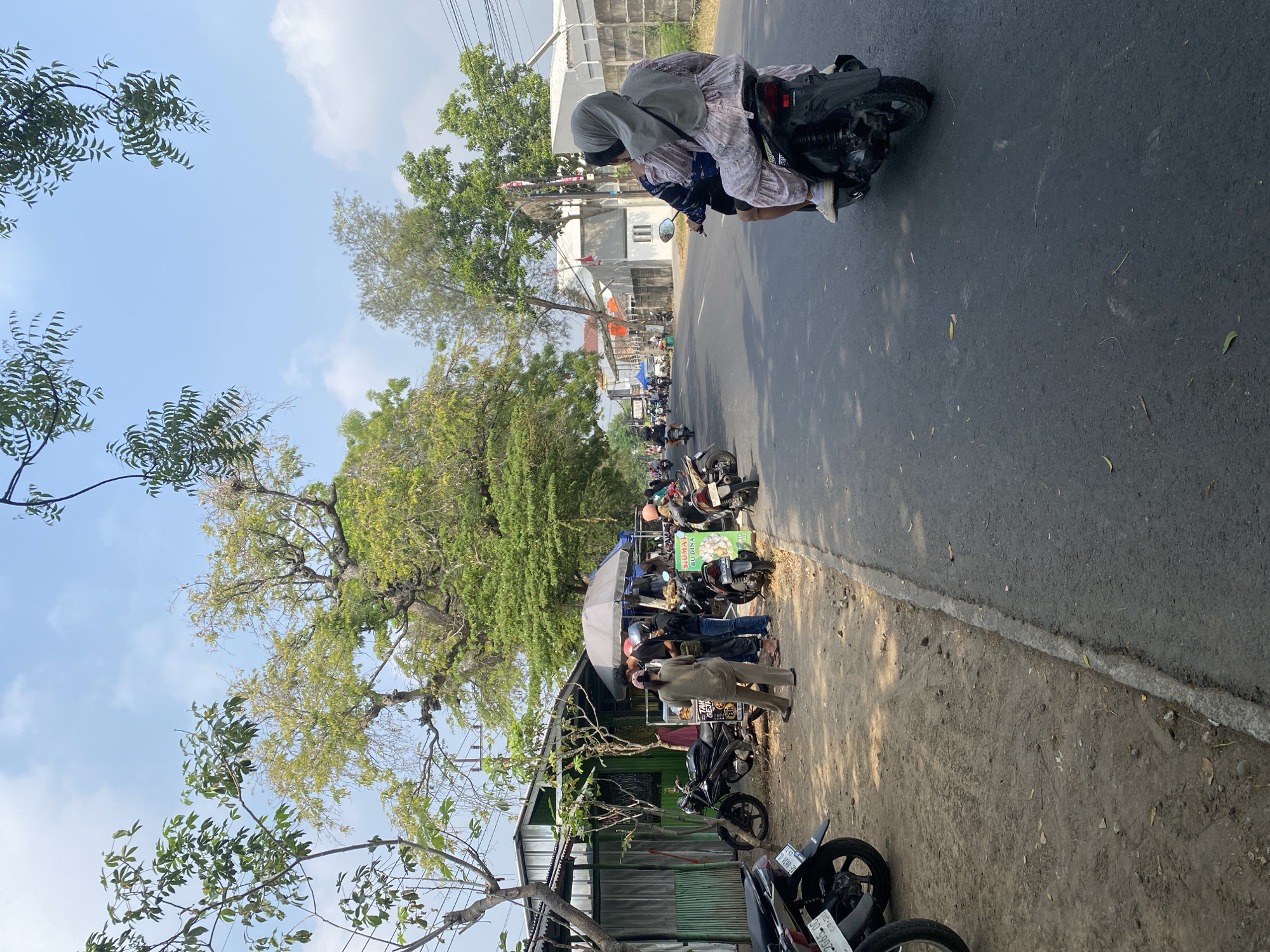 This is an eye-level, medium shot of a street scene in Banjarejo, Indonesia. The street is paved and lined with trees on both sides. On the left side of the street, there is a row of buildings with a dirt parking area in front. Several motorcycles are parked in the dirt area, and a few people are standing near a food stall with an umbrella. On the right side of the street, a motorcycle is driving away from the camera with two passengers. The sky is clear and blue with a few scattered clouds. The overall impression is a busy, everyday street scene in a tropical location.