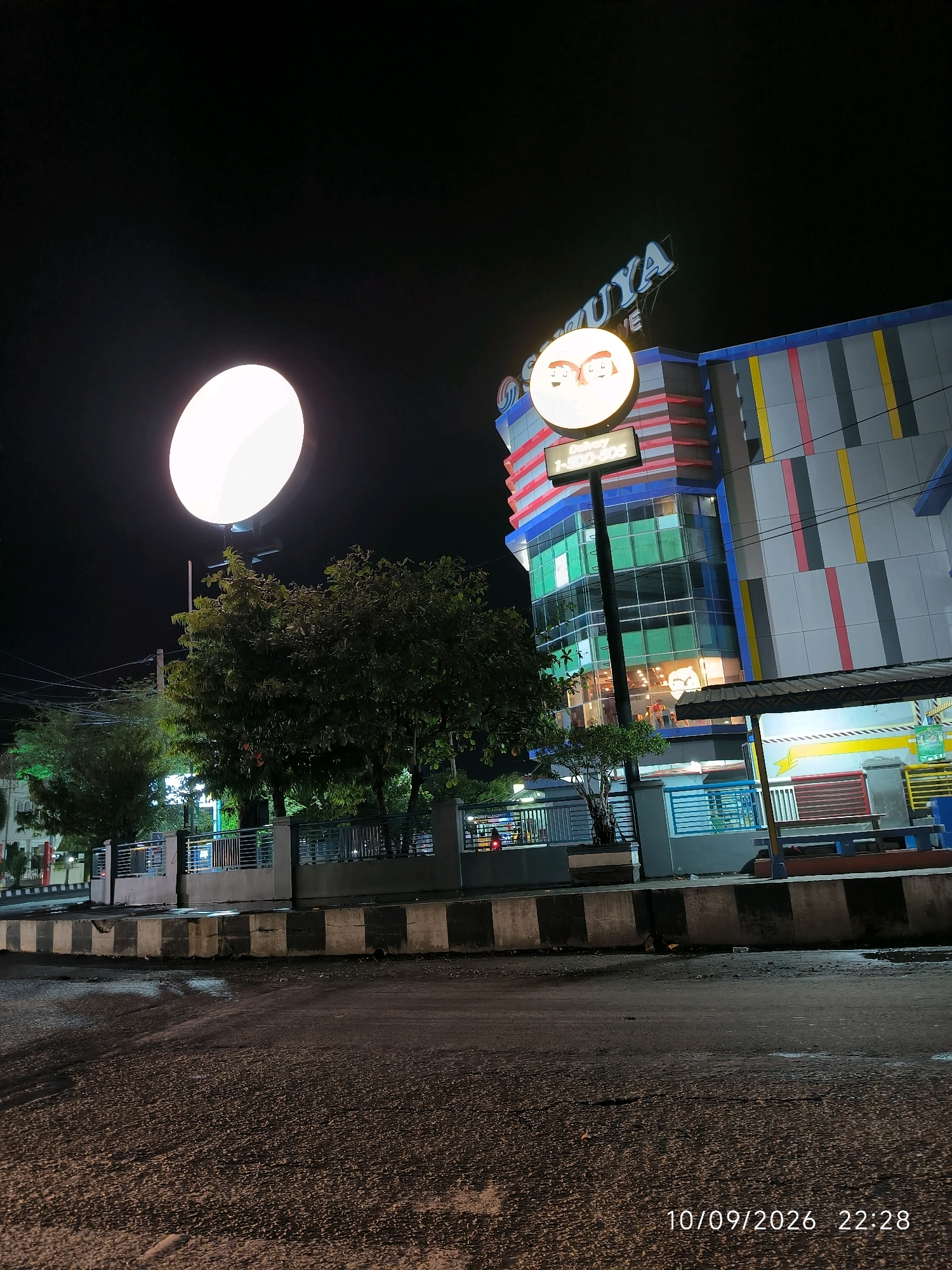 The image captures a nighttime view of a modern building complex in the city of Lhokseumawe, Indonesia. The building is illuminated by neon lights and has a striking architectural design with colorful vertical stripes. A large, circular illuminated sign with cartoonish eyes is prominently displayed near the top of the building. Below this sign, a smaller digital display shows the text "Delivery 1-800-505". The building appears to be a commercial establishment, possibly a shopping mall or a restaurant.
In the foreground, a paved road is visible, with a concrete divider separating it from a sidewalk area. A dark, mature tree with dense foliage stands to the left of the building, and other smaller trees and decorative fencing are also present. The sky is dark, indicating it is nighttime. The timestamp "10/09/2026 22:28" is overlaid on the bottom right corner of the image, suggesting the photo was taken on September 10, 2026, at 10:28 PM. There are no people visible in the image. The scene is well-lit by artificial lights, highlighting the building's features and the surrounding streetscape.