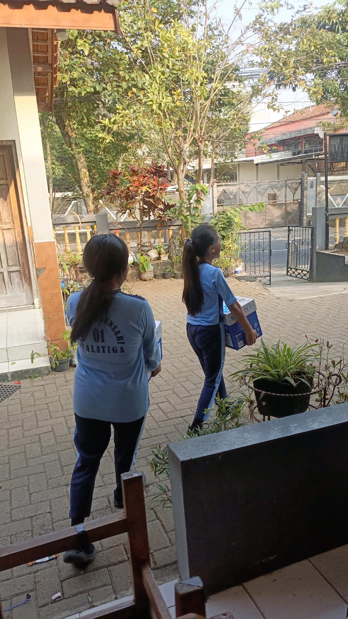 Two girls in light blue uniforms with dark blue pants are walking on a brick-paved walkway. The girl in the foreground has her back to the camera and is carrying a white box. The girl in the background is walking to the right and is also carrying a white box. Both girls have dark hair pulled back in ponytails. The environment appears to be outdoors, possibly at a school or educational institution in Salatiga, Indonesia, as indicated by the text on the back of the first girl's shirt which reads "GUNSARI 01 SALATIGA". There are trees and plants surrounding the walkway, and buildings can be seen in the background. The overall scene suggests a bright, sunny day.