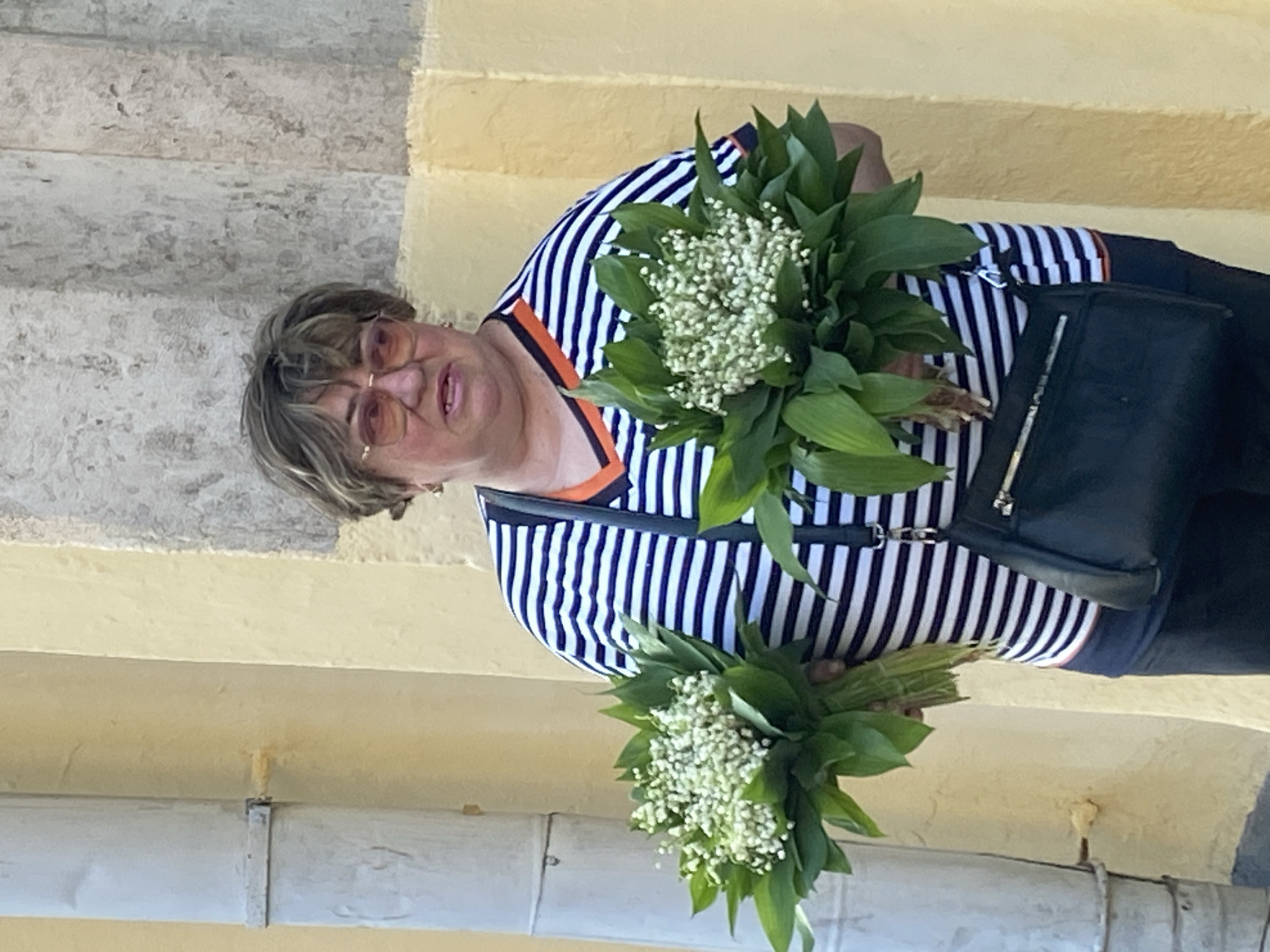 The image shows an older woman holding two bunches of Lily of the Valley flowers. She wears glasses and a striped shirt with orange trim, along with a dark-colored purse. The background suggests an urban environment, likely in Moscow, Russia, given the context and the presence of Lily of the Valley, a flower commonly sold on the streets there in spring. The woman stands near what appears to be a stone or concrete structure, possibly a building or staircase. The lighting suggests a sunny day.