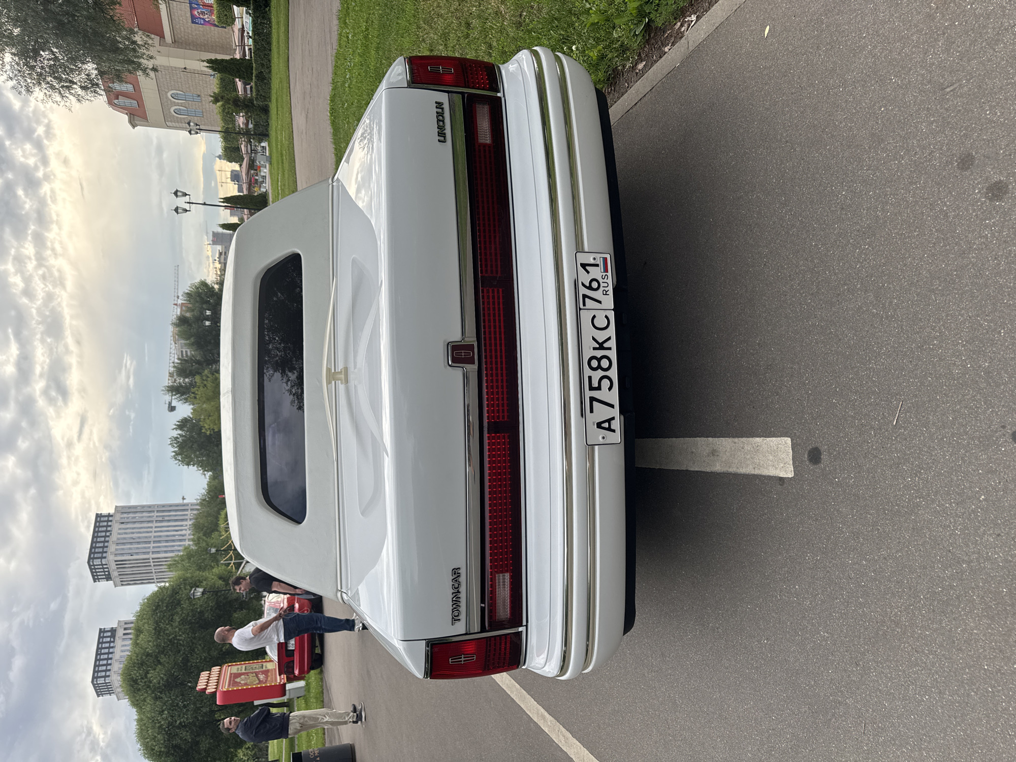 The rear view of a white Lincoln Town Car is captured on a paved surface, likely a parking lot or road, in a city environment. The car has chrome trim and a white vinyl roof, a common feature for limousines. The license plate, which is partially visible and reads "A758KC 761 RUS", indicates it is registered in Russia. In the background, a street with lamp posts, trees, and buildings can be seen. One building appears to be a shopping center or entertainment venue with an arched facade. In the left portion of the background, two men are standing near a red object with lights, possibly a booth or display. The sky is overcast, suggesting a cloudy day. The overall setting suggests an outdoor event or gathering in a public space in Moscow, Russia.