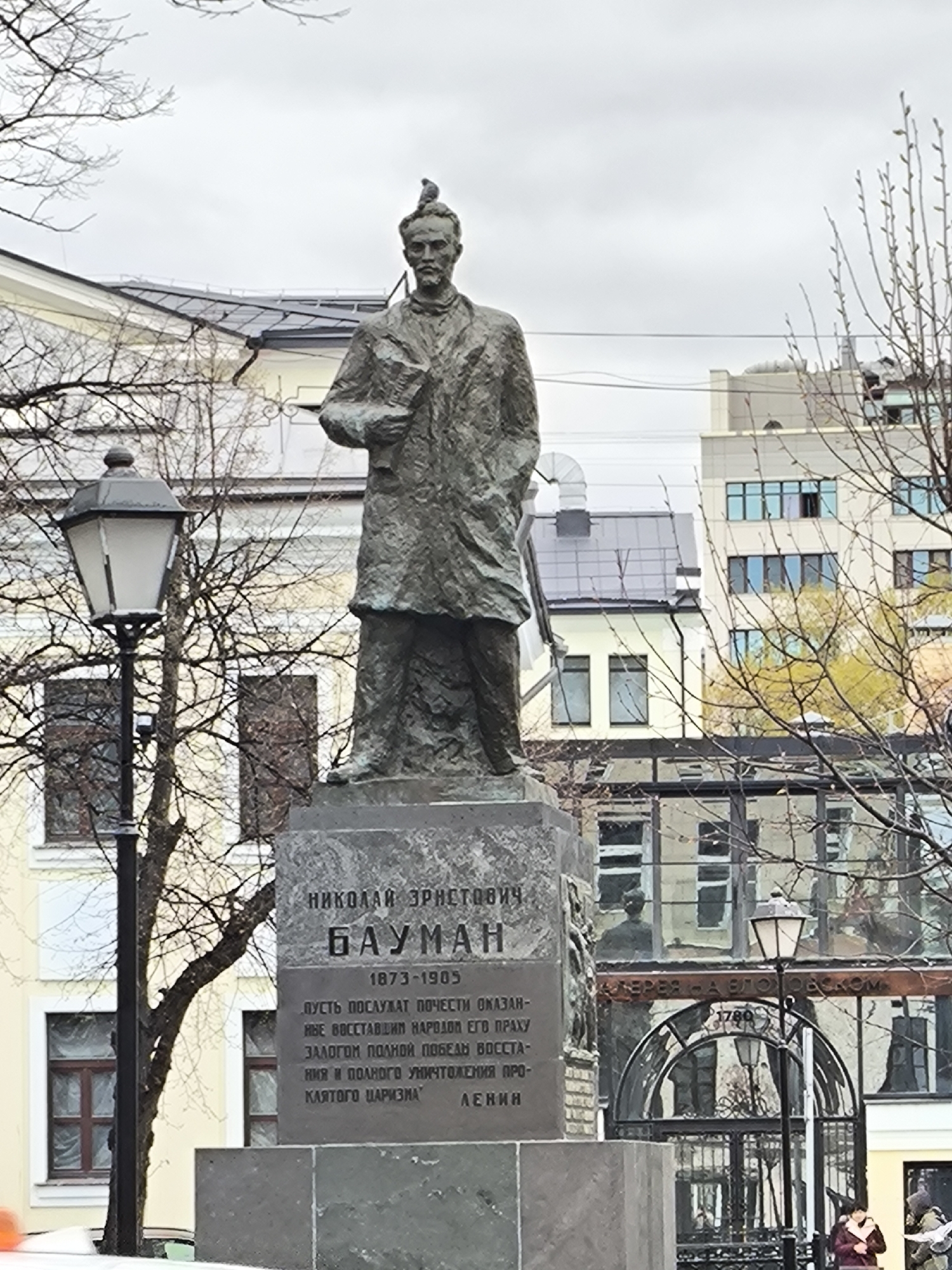 The image depicts the statue of Nikolai Bauman, a Russian revolutionary, in Moscow, Russia. The statue stands on a stone pedestal with Cyrillic inscriptions including his name, birth and death years (1873-1905), and a quote attributed to Lenin. A bird sits on the statue's head. Behind the statue, buildings with various architectural styles can be seen, with a mix of older and more modern structures. A street lamp is visible on the left, and bare branches of trees frame the scene. A metal gate is visible behind the statue. A person wearing a red coat is visible on the bottom right. The sky is overcast.
