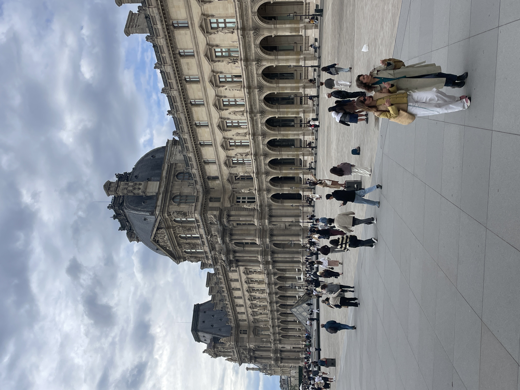 The image depicts the exterior of the Louvre Museum in Paris, France, under a cloudy sky. The grand architectural facade of the museum, characterized by its classical design, arched walkways, and ornate detailing, dominates the background. In the foreground, a large paved square is populated by numerous pedestrians walking in various directions. Among the people, two women are prominently featured in the lower right quadrant, walking together and engaged in conversation. The scene suggests a busy tourist day, with visitors exploring the iconic landmark.