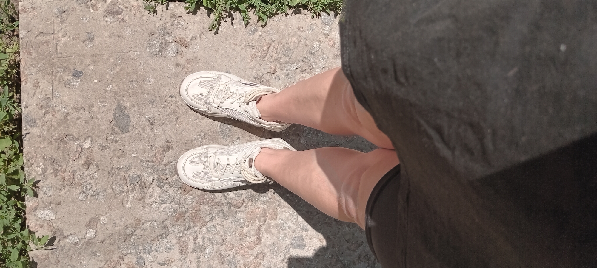 A first-person perspective shot shows a pair of white sneakers on a person's feet, standing on a concrete surface. The concrete has a rough, pebbled texture with visible aggregate. Green foliage is visible at the top and right edges of the frame, indicating an outdoor setting. Shadows are cast on the concrete, suggesting sunlight. The person is wearing black shorts. The image is taken in the city of Koviahy, Ukraine.