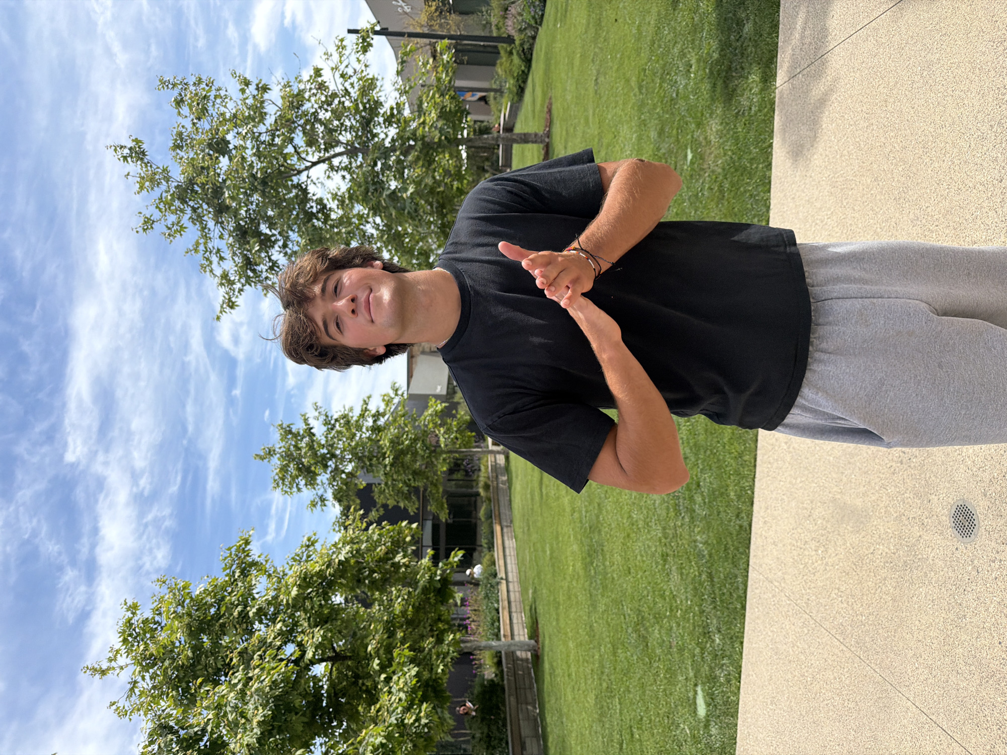 A young man stands on a beige paved surface next to a grassy area. He wears a black t-shirt and gray sweatpants, and has brown hair. He is looking at the camera with a smile and has his hands together in front of him. Behind him are trees and buildings under a blue, partly cloudy sky. The photo was taken in Malibu, California.