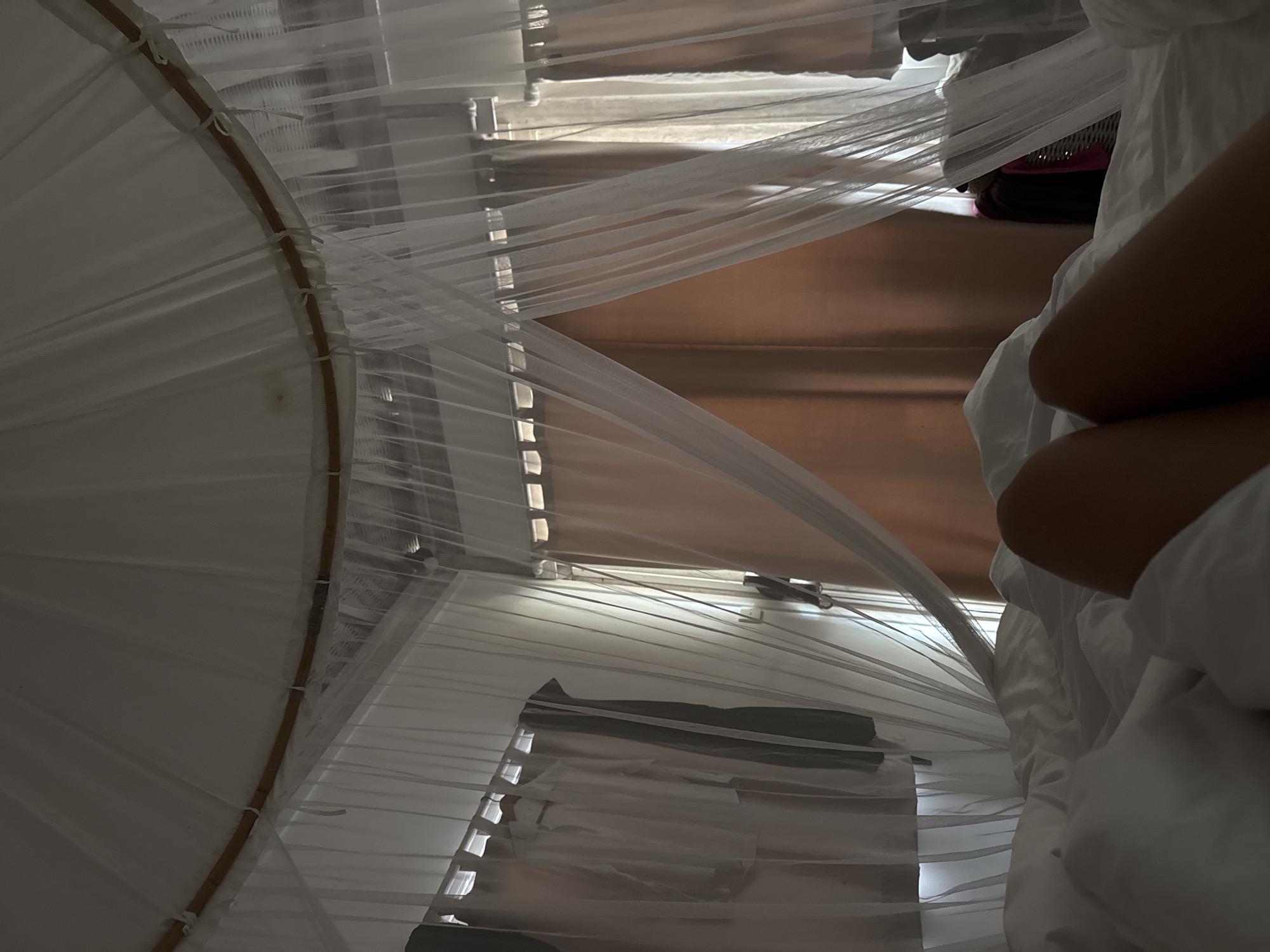 The image, taken from an upward angle, shows the interior of a room, likely in Unknown, Indonesia, possibly a bedroom, featuring a round mosquito net draped over a bed. The net is white and semi-transparent, obscuring much of the scene behind it. Through the net, one can see closed window blinds, indicating daylight filtering in. To the right of the image, a person's arm and part of their body are visible, covered by a white bedsheet, suggesting they are in bed. In the background, there is a glimpse of luggage, potentially a backpack, resting on the floor. The focus is on the bed, the net, and the presence of a person within the space.