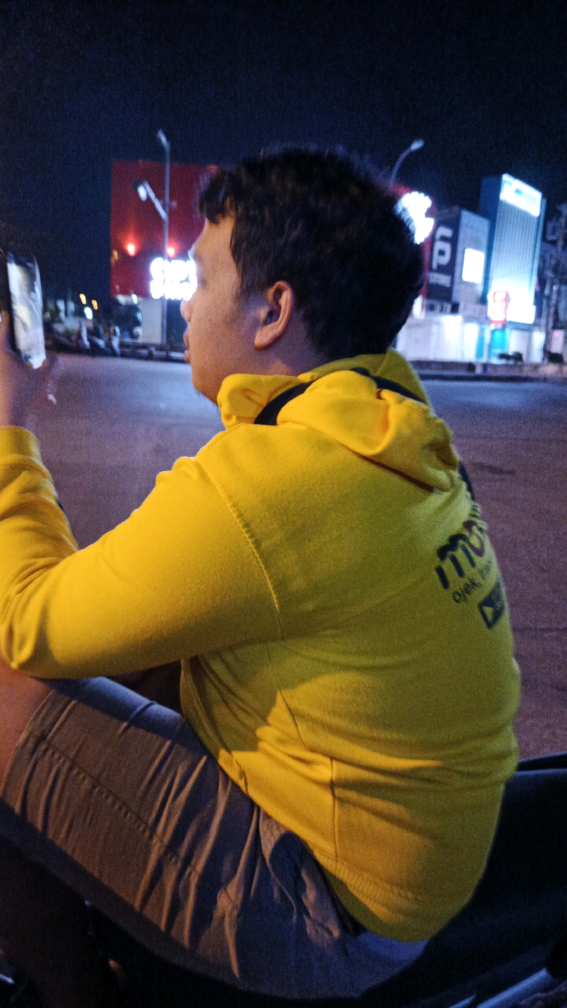 The image shows a person wearing a yellow hoodie and gray shorts, sitting outdoors at night. The person is holding a smartphone in their left hand and looking to the right. In the background, there are illuminated buildings and streetlights, suggesting an urban environment. The text on the hoodie is partially visible and appears to be in a dark color. The lighting and setting indicate this scene is likely in a city, and the provided information confirms the location as Surakarta, Indonesia. The image is captured in a slightly grainy, low-light condition.