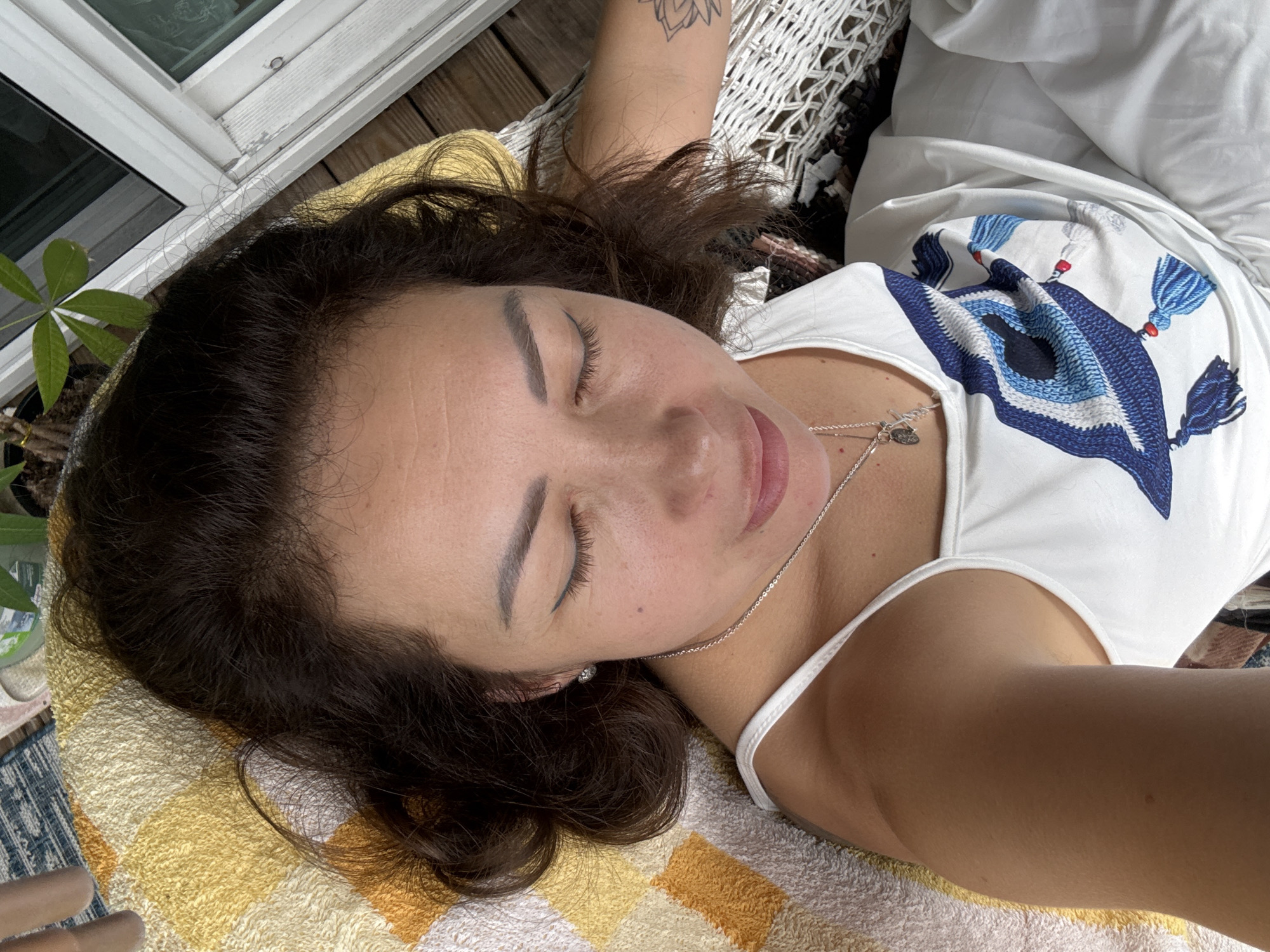 The image shows a close-up, overhead view of a woman lying down with her eyes closed. She has dark, wavy hair that falls around her face and shoulders. She is wearing a white tank top with a blue and white evil eye design on it, and a delicate silver necklace. Her skin appears to be tan. She is lying on a yellow and white checkered towel, which is on a wooden deck. To the left of the woman, part of a white window frame and some green foliage are visible, suggesting an outdoor setting. A portion of a white hammock with a net-like pattern is also visible behind her head. The lighting suggests it is daytime, possibly a sunny day, given the bright illumination. The overall mood conveyed by the image is one of relaxation and peace. There is no visible text in the image.