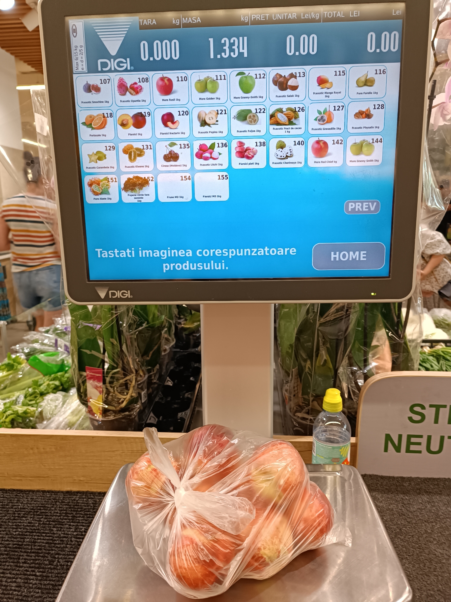 The image shows a close-up of a digital scale display in a supermarket. The display shows product information, prices, and weight, with a large number "0.000" indicating zero weight. The scale is in focus, while the background is blurred, showing a person in a striped shirt and denim shorts, and produce in plastic packaging. The setting is likely a self-checkout area in a grocery store in Chișinău, Moldova, as suggested by the Romanian language on the screen and the common retail equipment. The scene captures a moment of weighing produce, with a bag of apples prominently displayed on the scale.