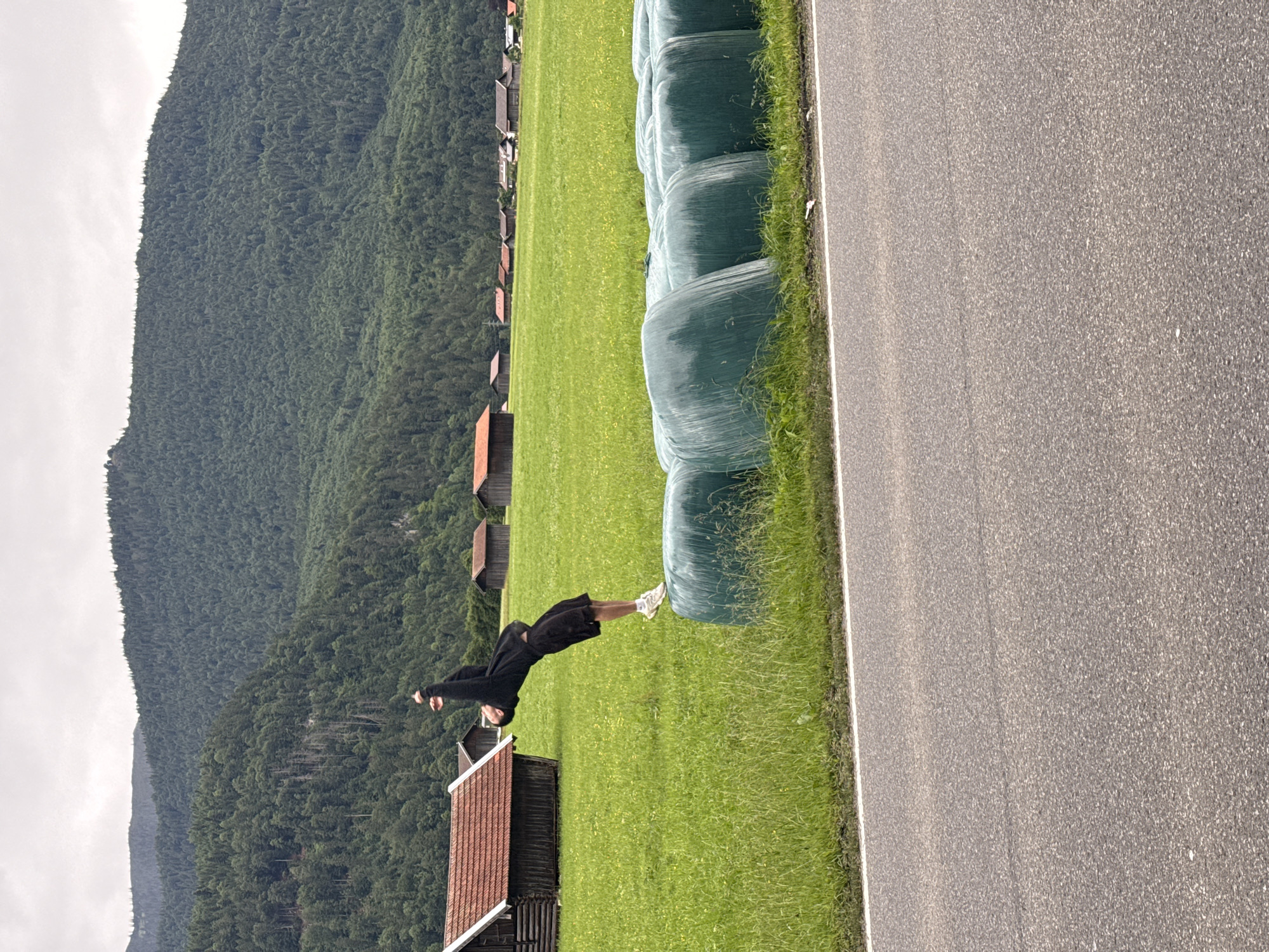 In a pastoral landscape near Garmisch-Partenkirchen, Germany, a young individual in black shorts and a black hoodie is captured mid-flip. They are performing an acrobatic maneuver, leaping over a row of large, green-wrapped hay bales that line the edge of a vibrant green field. The field, dotted with yellow wildflowers, stretches towards a backdrop of traditional Bavarian buildings with red roofs and wooden structures. Beyond the buildings, a dense, dark green forest covers the lower slopes of a majestic mountain range. The sky above is overcast, casting a diffused light on the scene. A paved road runs across the foreground of the image, separating the viewer from the field and the acrobatic action.