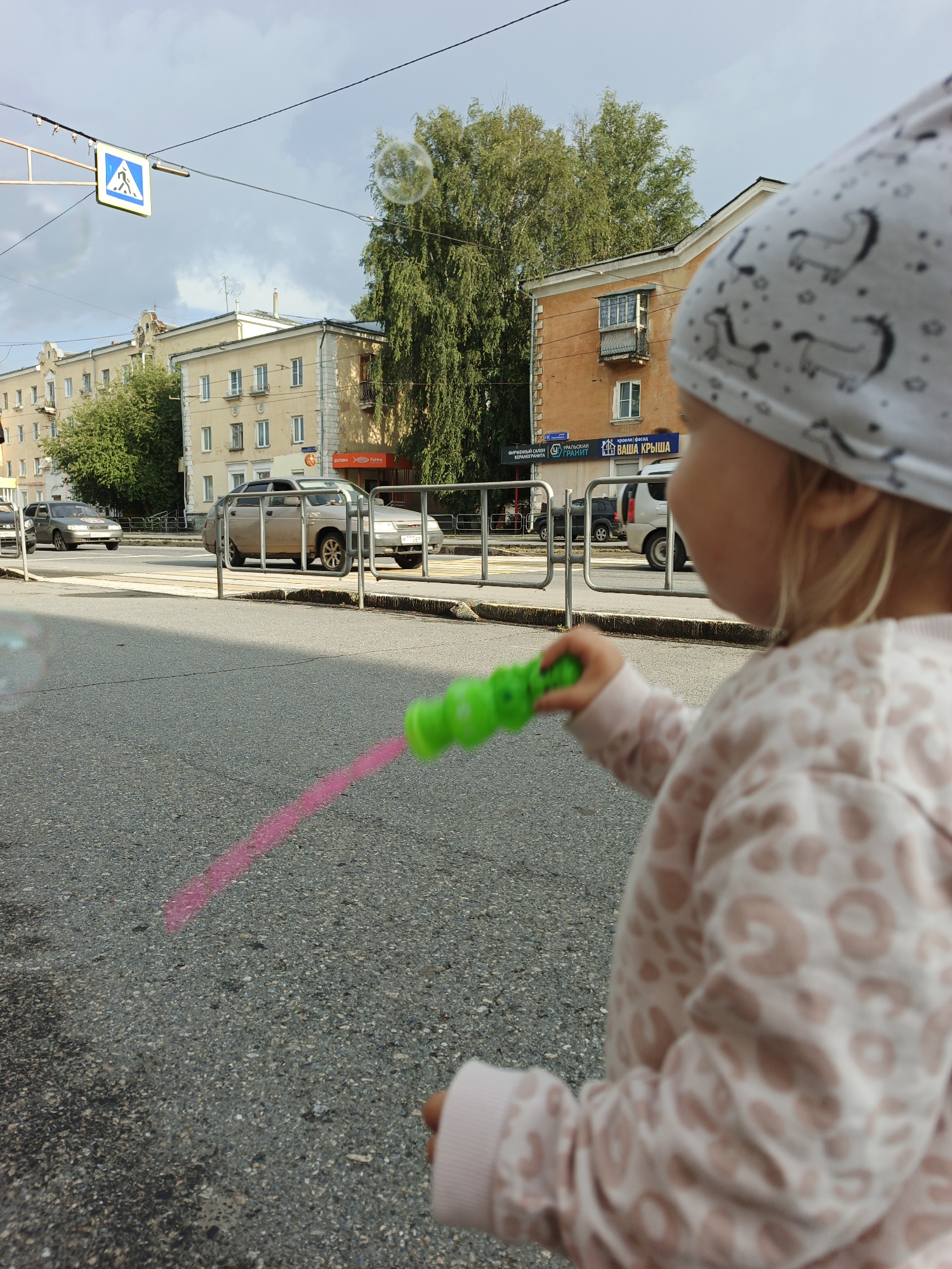 The image depicts a young child, facing away from the camera, holding a green bubble wand with a pink trail of soap suds, seemingly blowing bubbles on a street in Zlatoust, Russia. The child is wearing a light pink hooded jacket with a leopard-like pattern and a white beanie adorned with small animal figures. In the background, a street with several cars, including a silver sedan and a silver SUV, is visible. Across the street, there are multi-story buildings with cream and orange facades, some with balconies and windows. Trees line the sidewalk in front of the buildings. A pedestrian crossing sign is posted on the left side of the image. The sky is overcast with clouds.