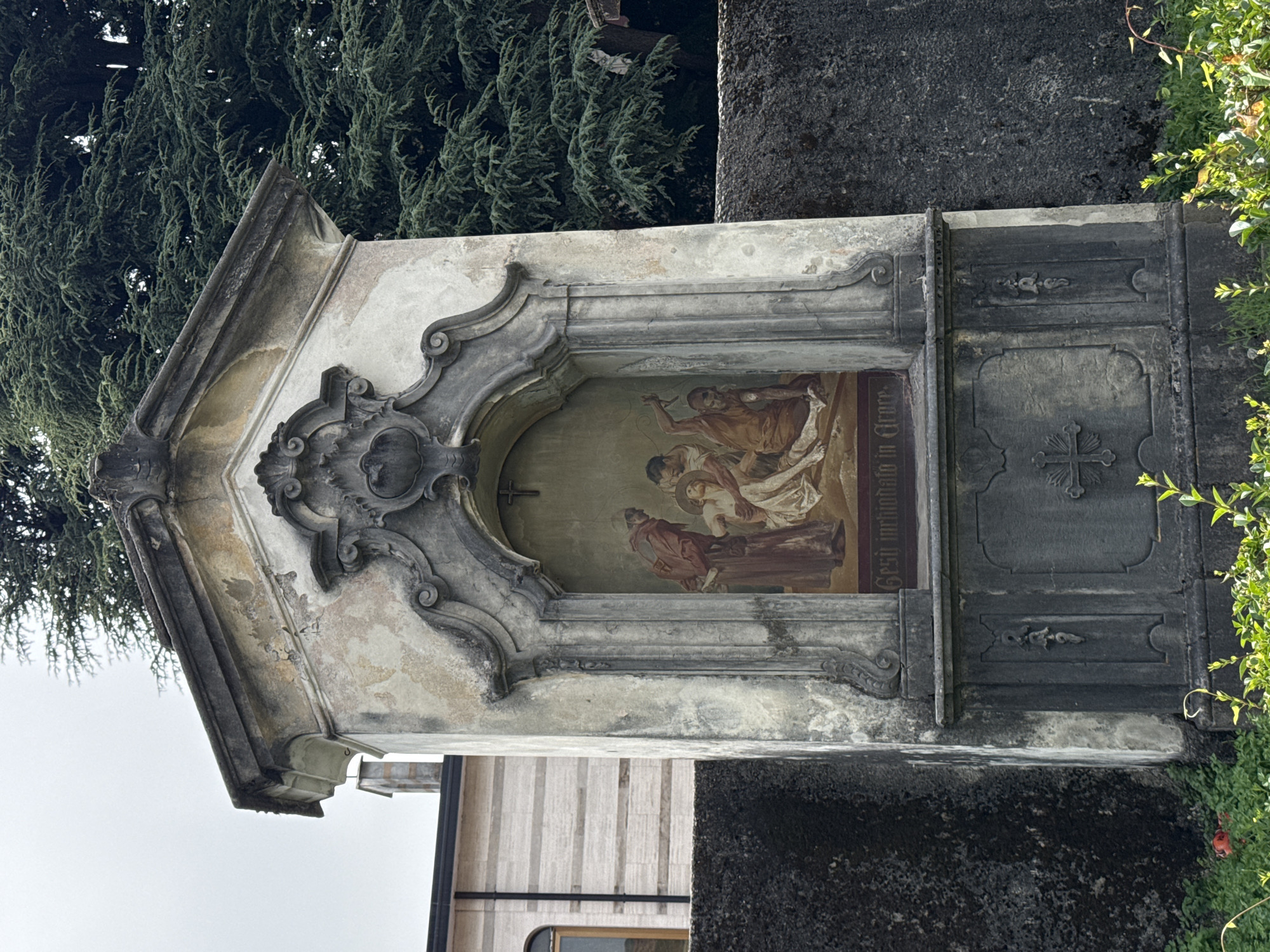 This is a medium shot, eye-level outdoor photograph of a weathered, stone wayside shrine situated in front of a dark stone wall. The shrine is a vertical structure with a pitched roof. The main body of the shrine has an arched niche in the center, which contains a painting depicting a religious scene. The painting shows figures in robes, with one figure appearing to hold another in their arms, and another figure is seated on the ground, possibly hammering. The inscription "Gesù inchiodato in Croce" is visible below the painting. The shrine is decorated with ornate stonework and shows signs of age and deterioration, including peeling paint and moss growth. Below the niche, there is a darker stone section with decorative carvings, including a cross on the left and a bell-shaped ornament on the right. In the foreground, there is lush green vegetation, with a single red flower visible on the left. In the background, a large evergreen tree with dense foliage dominates the right side of the frame. To the left of the shrine, a modern building with horizontal siding and a window is partially visible. The sky is overcast.