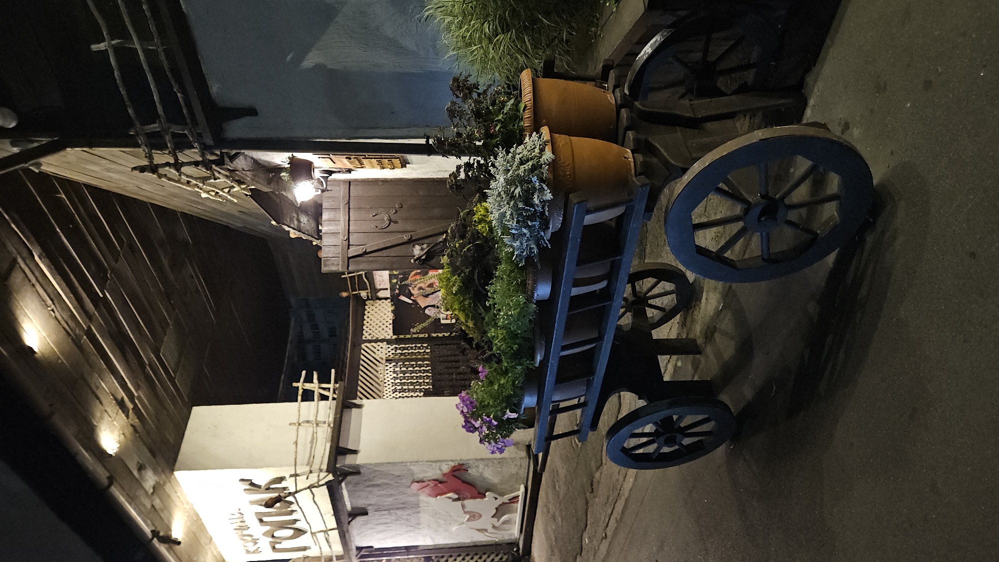 This image depicts a nighttime scene outside a building with Cyrillic signage that reads "КОРЧМА ГОРЯК". The building has a rustic facade with wooden elements and is illuminated by overhead lights. In the foreground, a dark-colored wooden cart with four spoked wheels is filled with potted plants. The plants include various green foliage and some flowering plants with purple blossoms. The cart is positioned on an asphalt surface, casting shadows. Behind the cart, the building features a large wooden door with a horseshoe emblem and a hanging lantern. The overall atmosphere is that of an outdoor dining or entertainment venue with a traditional or rustic theme. The text on the building, "КОРЧМА ГОРЯК", suggests it is a tavern or inn. While the image does not explicitly state the location, "КОРЧМА" is a common term for taverns in Slavic countries, and "ГОРЯК" could be a proper name or refer to a hot beverage. Without additional context or specific identifying markers within the image, pinpointing the exact location within Ulyanovsk, Russia is not possible.