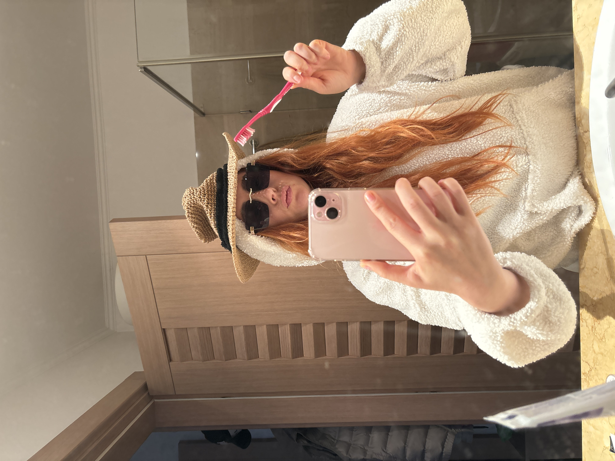A person with long red hair wearing sunglasses and a straw hat is taking a selfie in a bathroom mirror. They are holding a pink toothbrush and a light pink smartphone. The bathroom has wooden cabinets and a shower with glass doors. Based on the style and features of the bathroom, and the image's origin, the location is likely in Budva, Montenegro, though this cannot be forensically confirmed from the image alone.