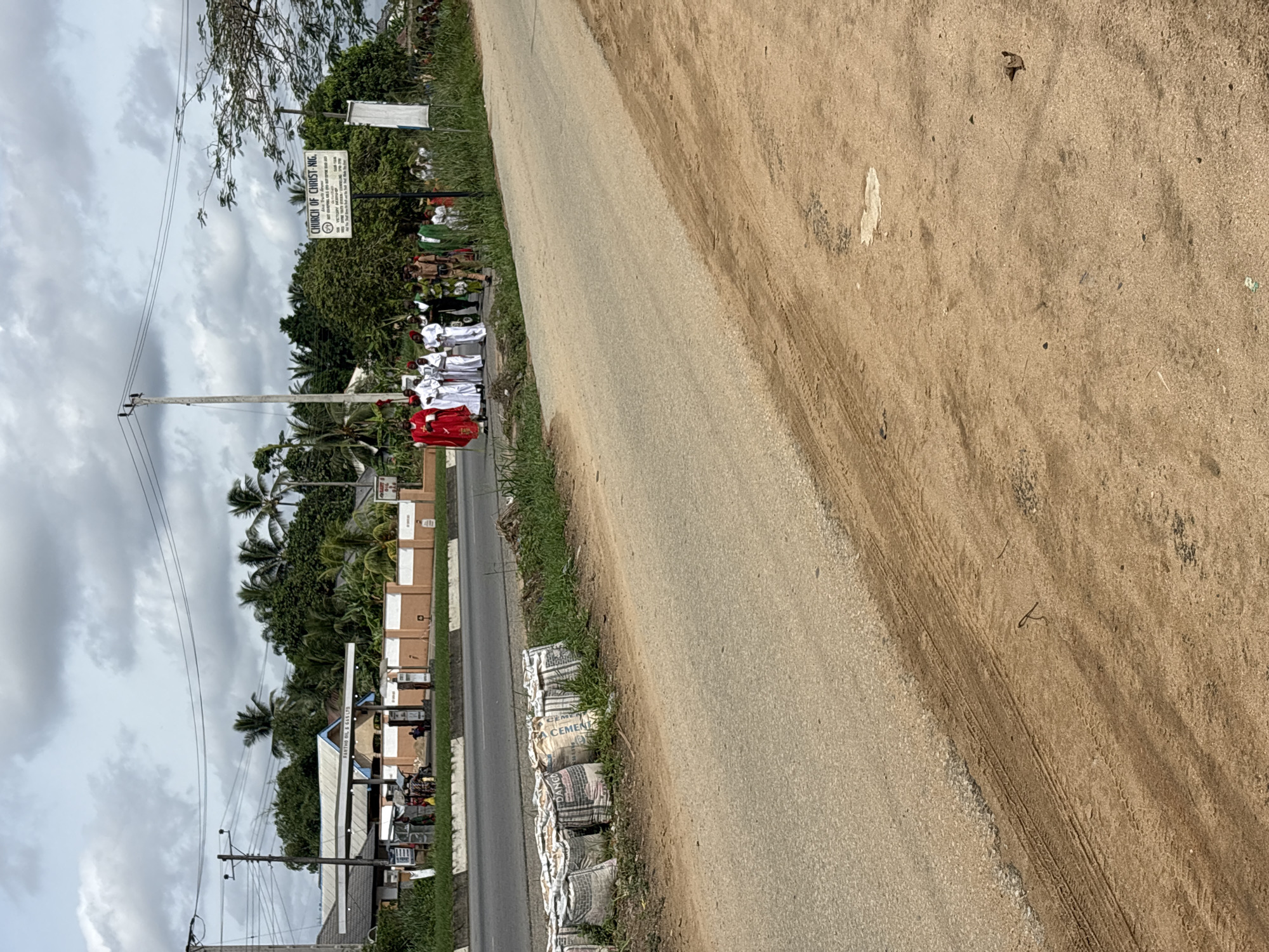The image depicts a road scene in Use Ikot Usuan, Nigeria. A group of individuals in religious attire, including red and white robes, are walking on the shoulder of a paved road lined with grass. Adjacent to the road is an unpaved area of dirt. In the background, there's a gas station, utility poles, and dense vegetation, including palm trees. A sign for a church is also visible. Cement bags are stacked along the side of the road closer to the gas station. The sky is overcast.