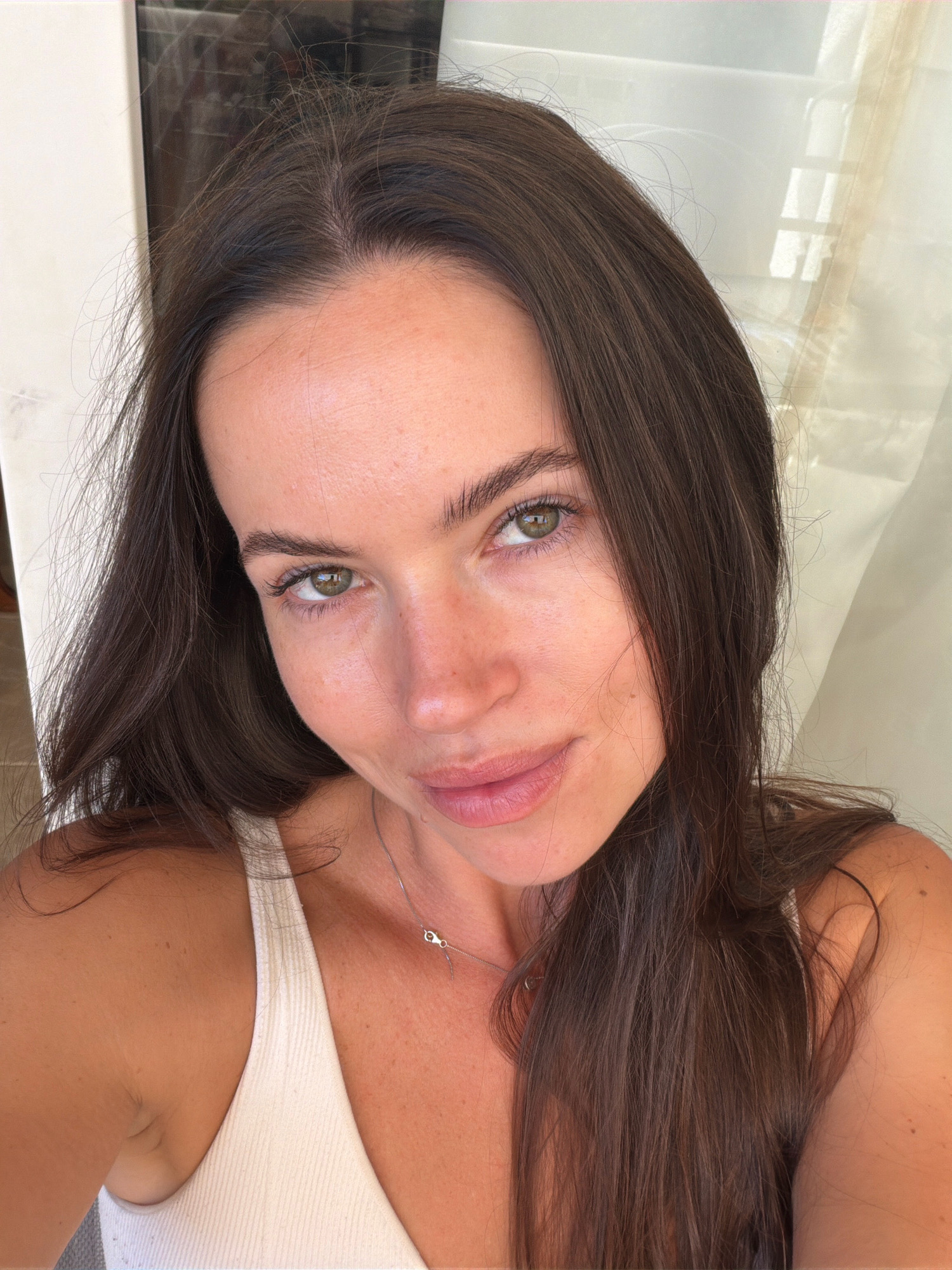A woman with long, dark brown hair and green eyes is captured in a close-up selfie. She is wearing a white, ribbed tank top and a delicate silver necklace with a small pendant. The background features a light-colored curtain with window-like patterns visible through it, and a hint of what appears to be a glass door on the left side. The lighting suggests an indoor setting, possibly with natural light filtering through the curtain. No specific location in Salou, Spain, is identifiable from the image alone.