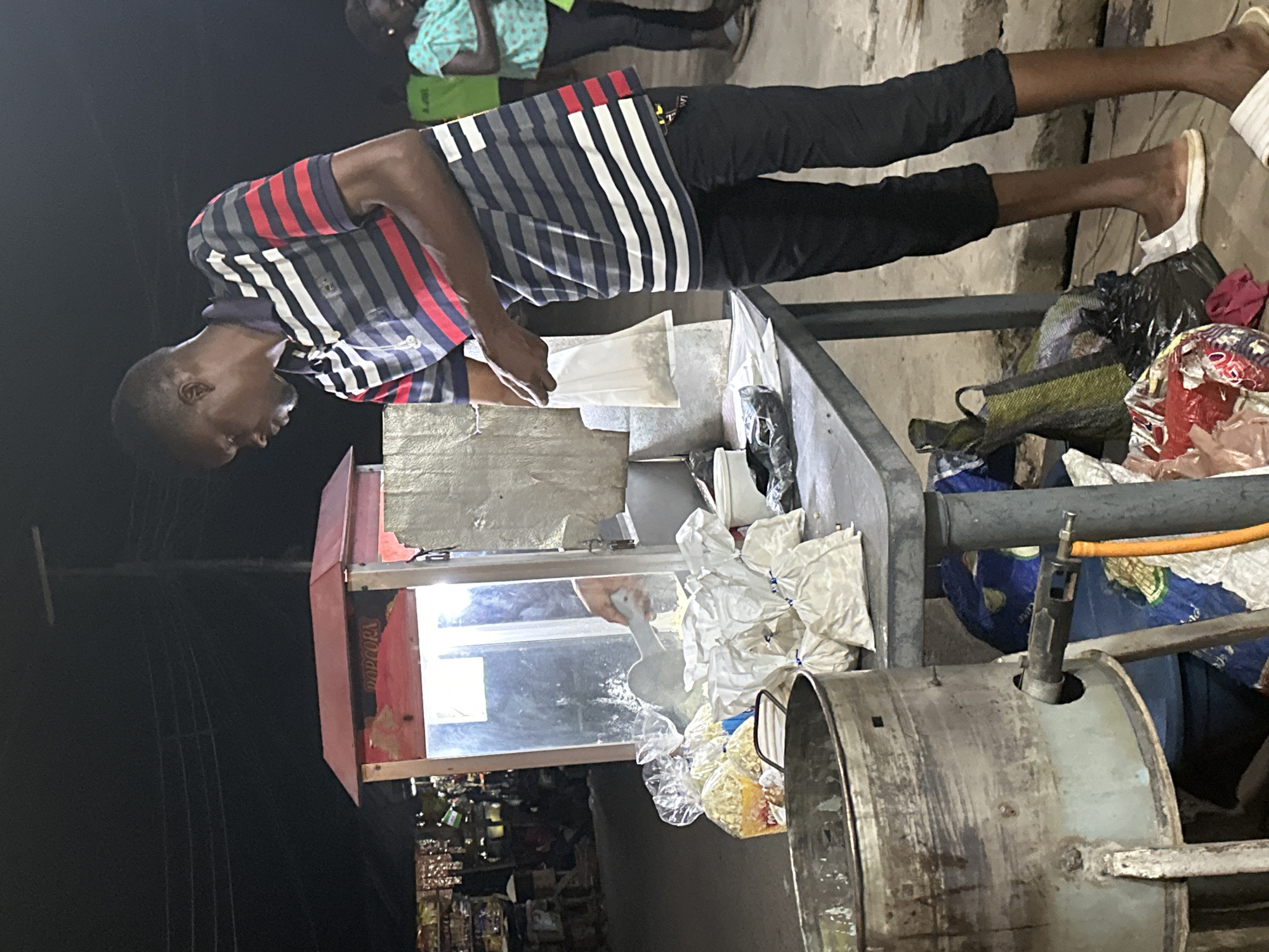 At night in an urban location in Unknown, Unknown, a man in a striped shirt and black pants stands beside a metal food vending cart illuminated by an overhead light. He is filling a small plastic bag with a food item, likely popcorn, from a container on the cart. Various bags and containers surround the cart, and a metal cooking apparatus is visible on the right side. In the background, a dimly lit storefront displays a variety of goods. Other people are present in the background.