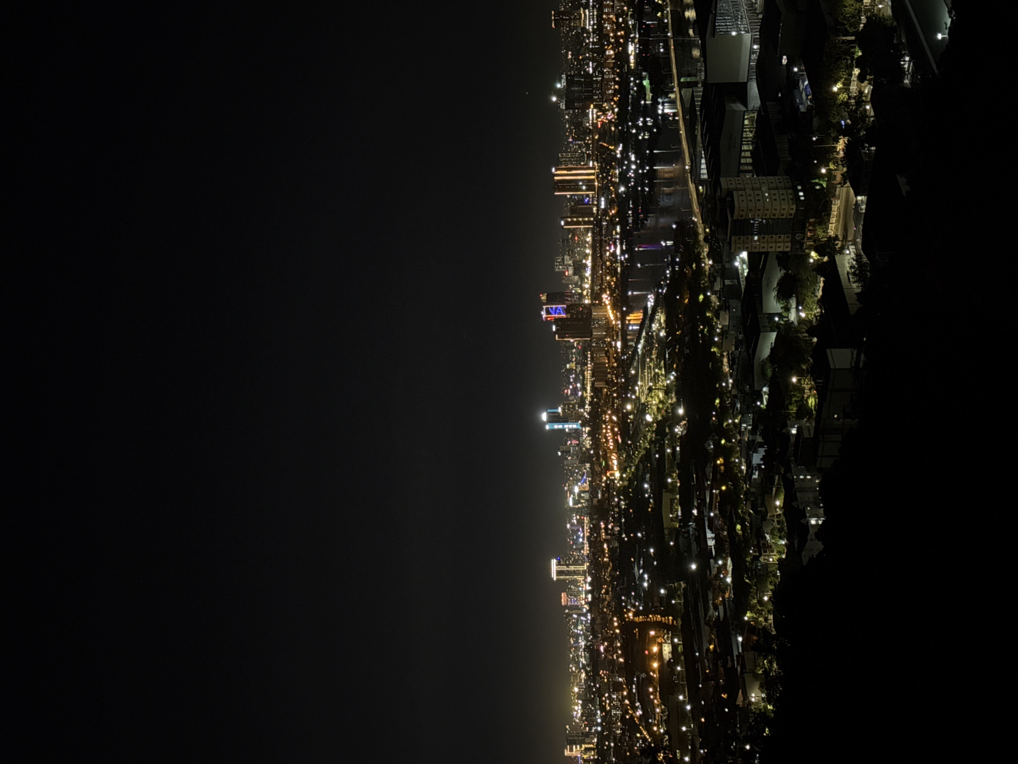 This is an aerial night-time photograph of the city of Đà Nẵng, Vietnam, viewed from a high vantage point, likely a hill or mountain overlooking the city. The foreground is dominated by a dark, silhouetted landscape, possibly foliage and the crest of the hill from which the photo was taken.
The middle ground and background are a vibrant tapestry of city lights. A prominent feature is the Han River, which winds through the city, its dark waters reflecting the bright lights of buildings and bridges. The river is crossed by several bridges, visible as illuminated structures.
On either side of the river, the city sprawls with countless lights from buildings, streets, and residential areas. Several modern skyscrapers with distinctive lighting patterns stand out in the skyline. Some buildings are illuminated with warm yellow or orange lights, while others feature cool blue or white lighting. The overall impression is a bustling and dynamic urban environment at night.
The sky is a deep, inky black, typical of a night scene with minimal ambient light pollution. There are no visible people or specific actions taking place, and the image focuses solely on the urban landscape and its illumination. The composition creates a sense of depth, with the dark foreground contrasting sharply with the brightly lit city.